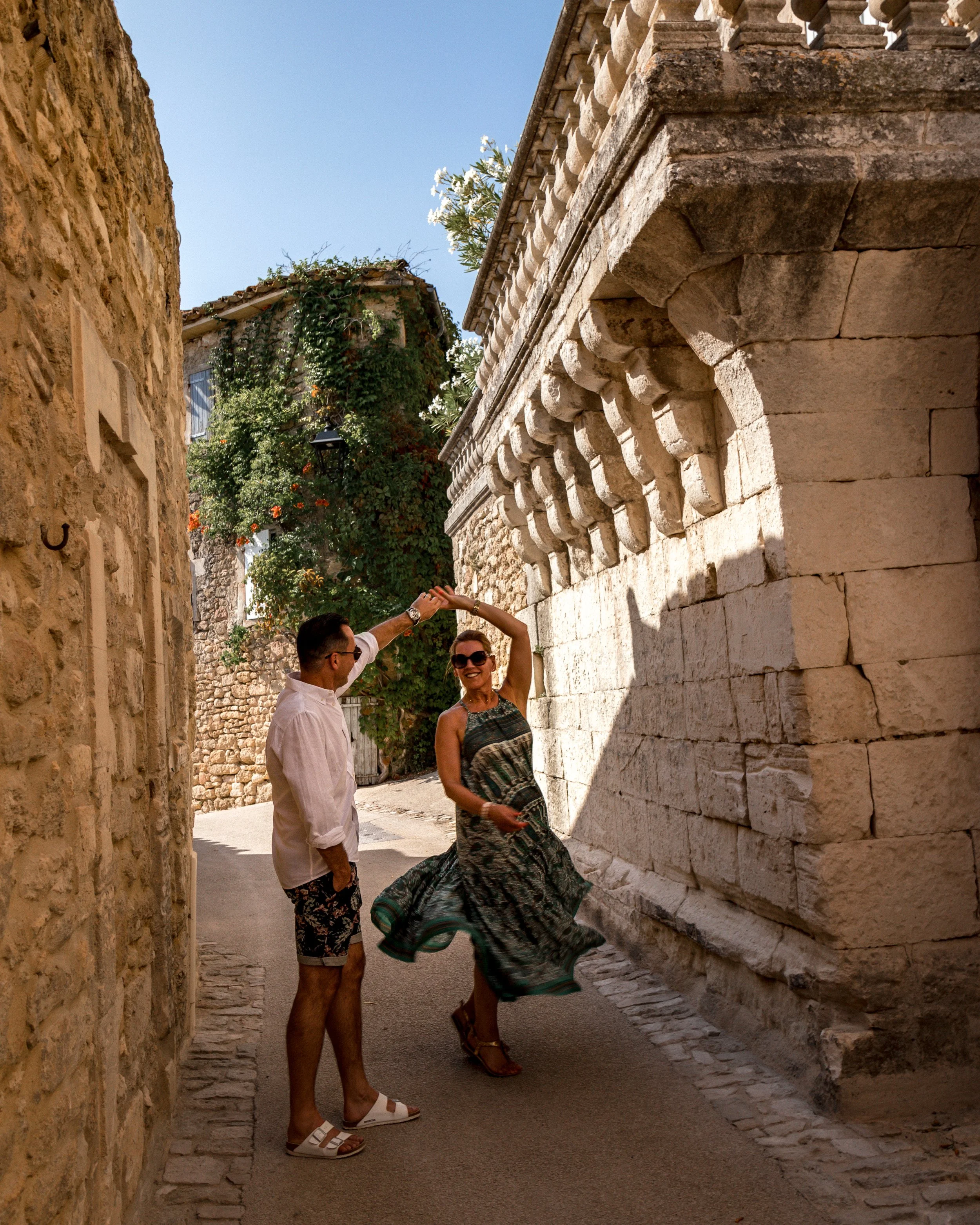 A man and woman dance together on a narrow street with stone buildings, with the man holding the woman's hand as she twirls.
