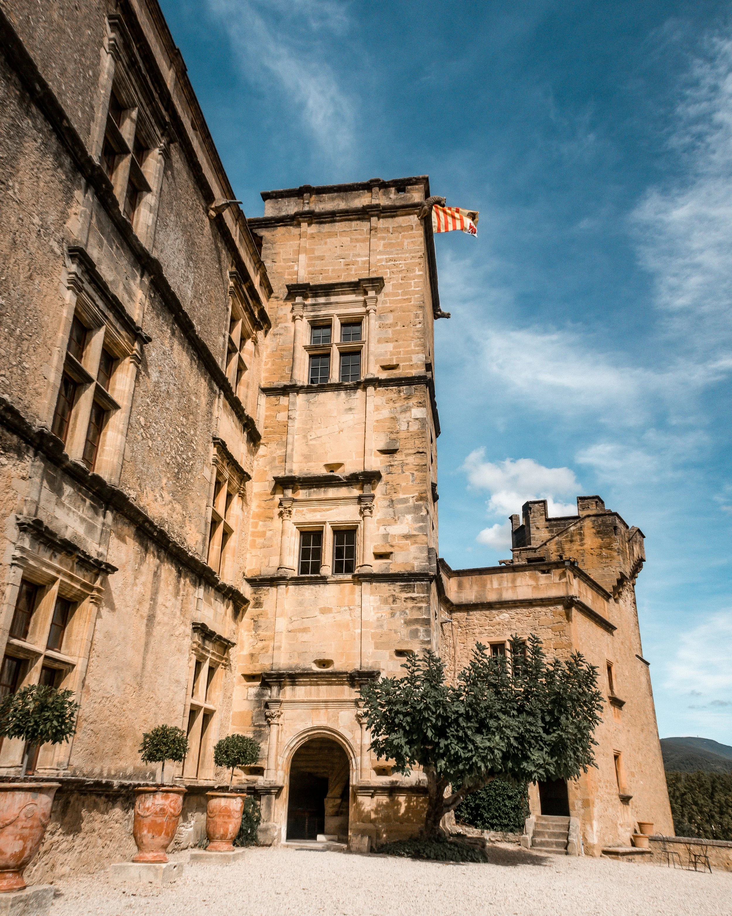A historic castle with stone walls, multiple windows, and a flag on a pole in the top corner, set against a blue sky with clouds, surrounded by small trees and gravel ground.