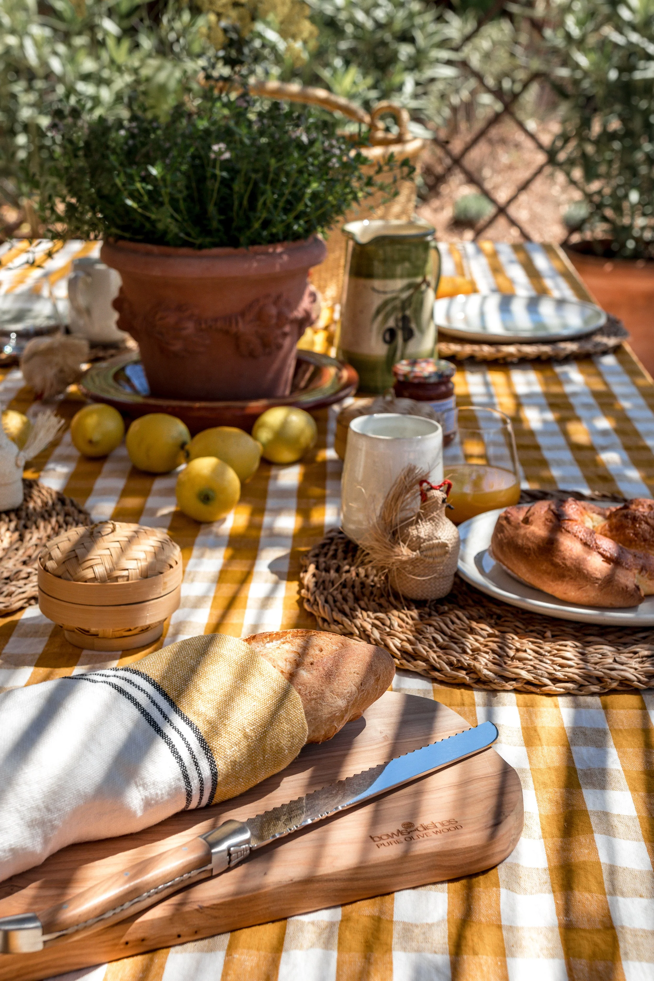 Outdoor table set for a meal with a yellow and white checkered tablecloth, bread, apples, glasses of beverage, a potted plant, and dishes.