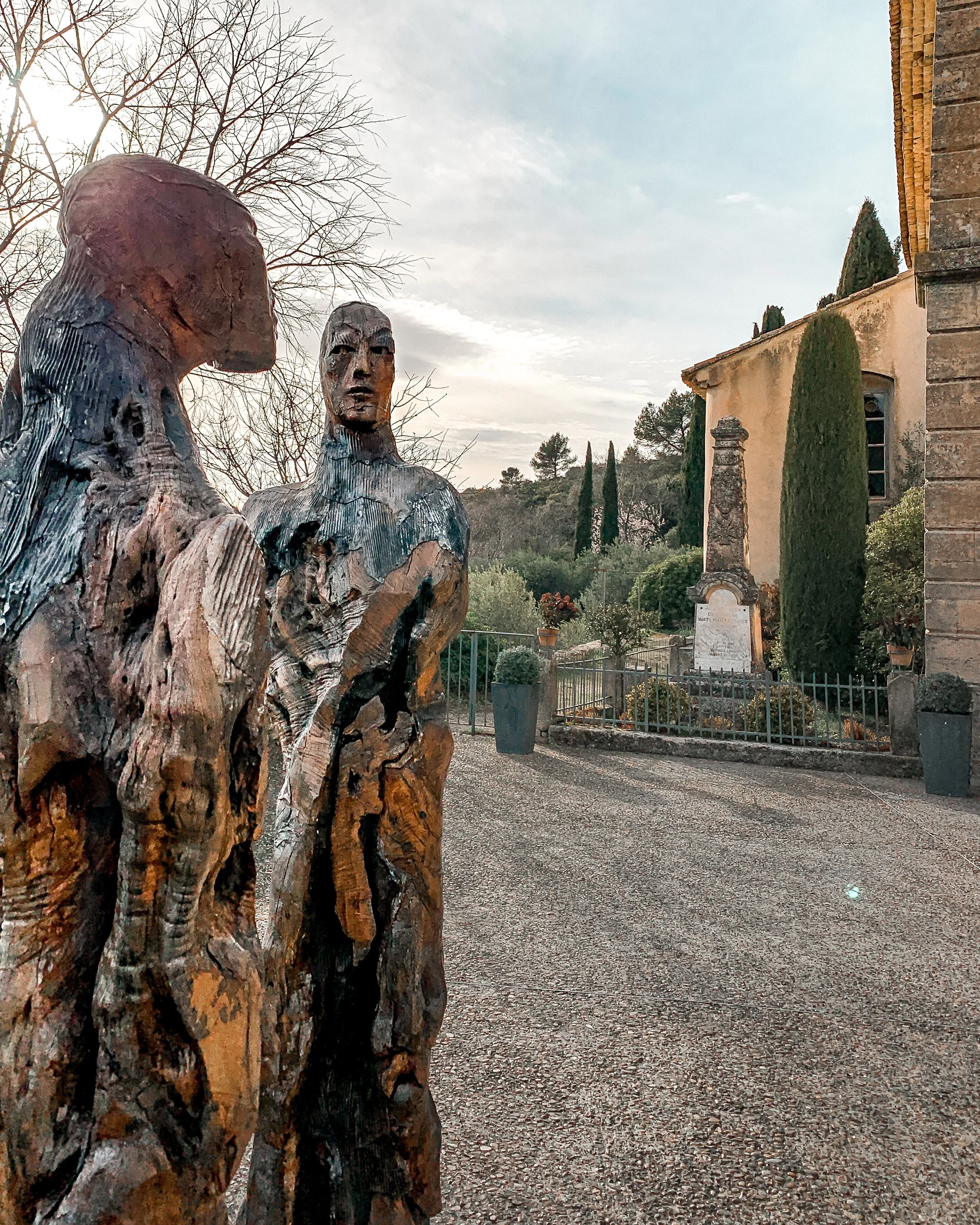 Wooden sculptures of two human figures in the foreground with a historic building, trees, and a garden in the background.