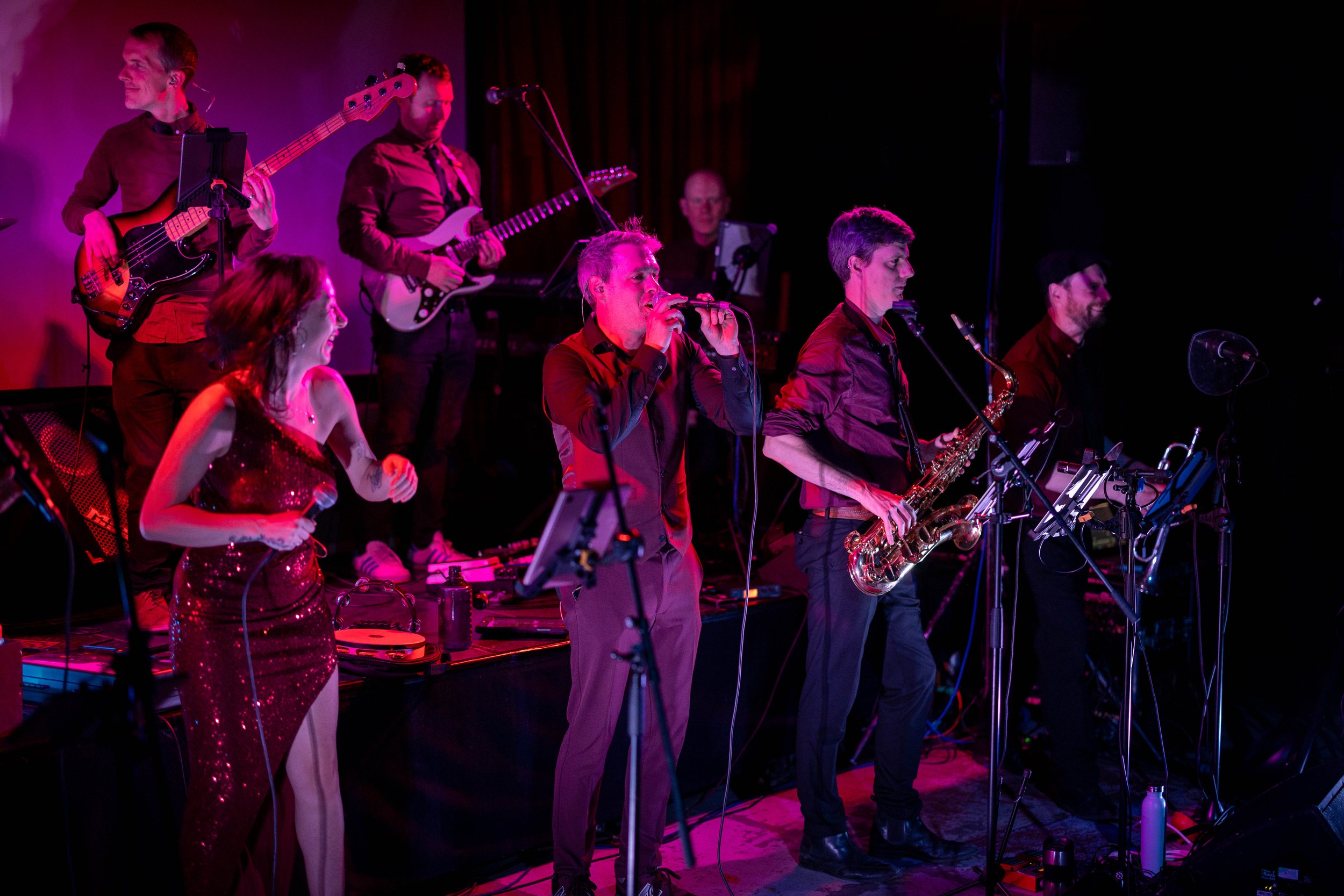 Hot Jazz Biscuits performing on stage with vocalists, guitars, saxophone and drums under purple lighting.