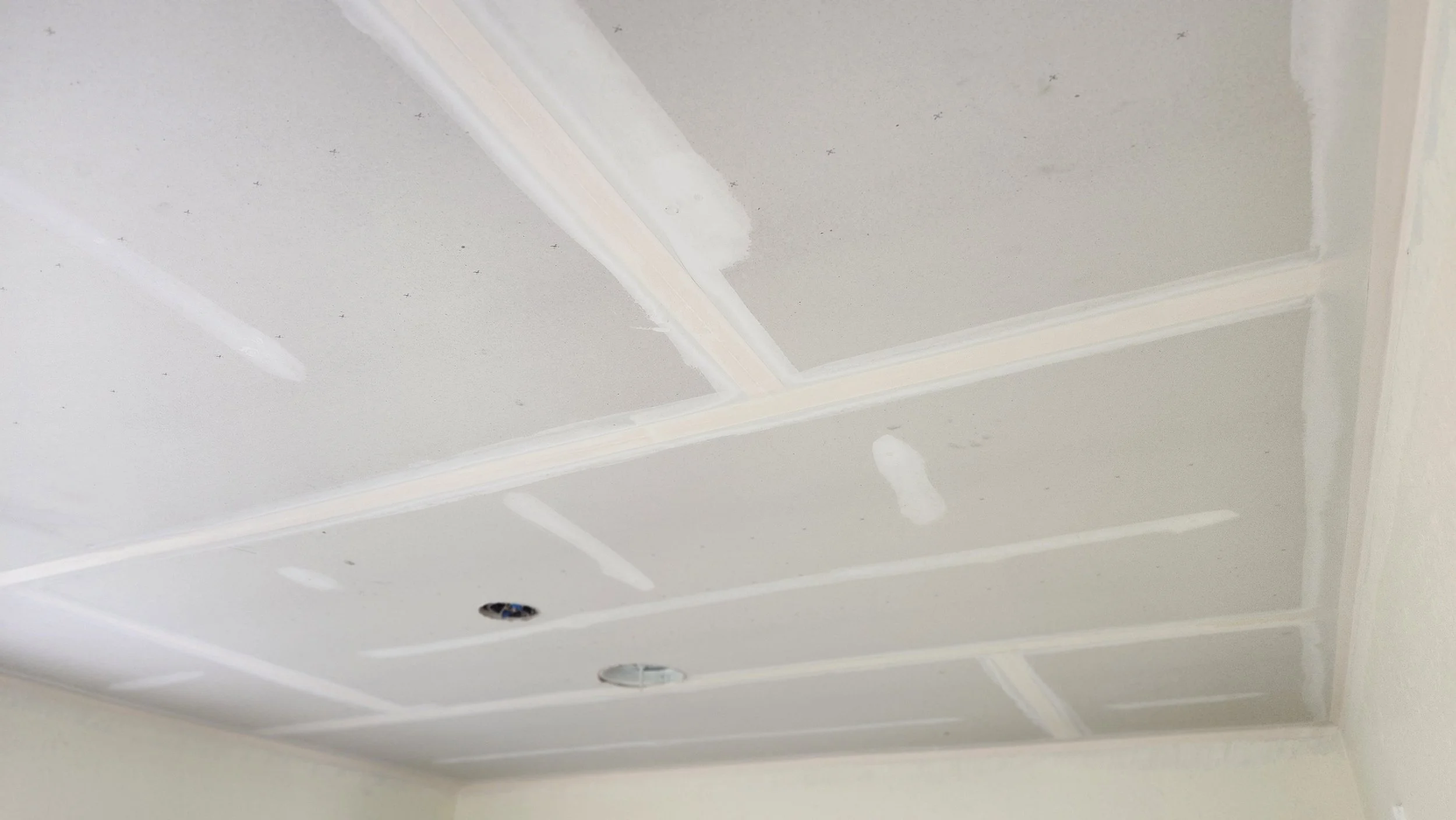 Ceiling with drywall panels installed and joint compound applied to seams, with one electrical fixture hole open, awaiting light fixture installation.