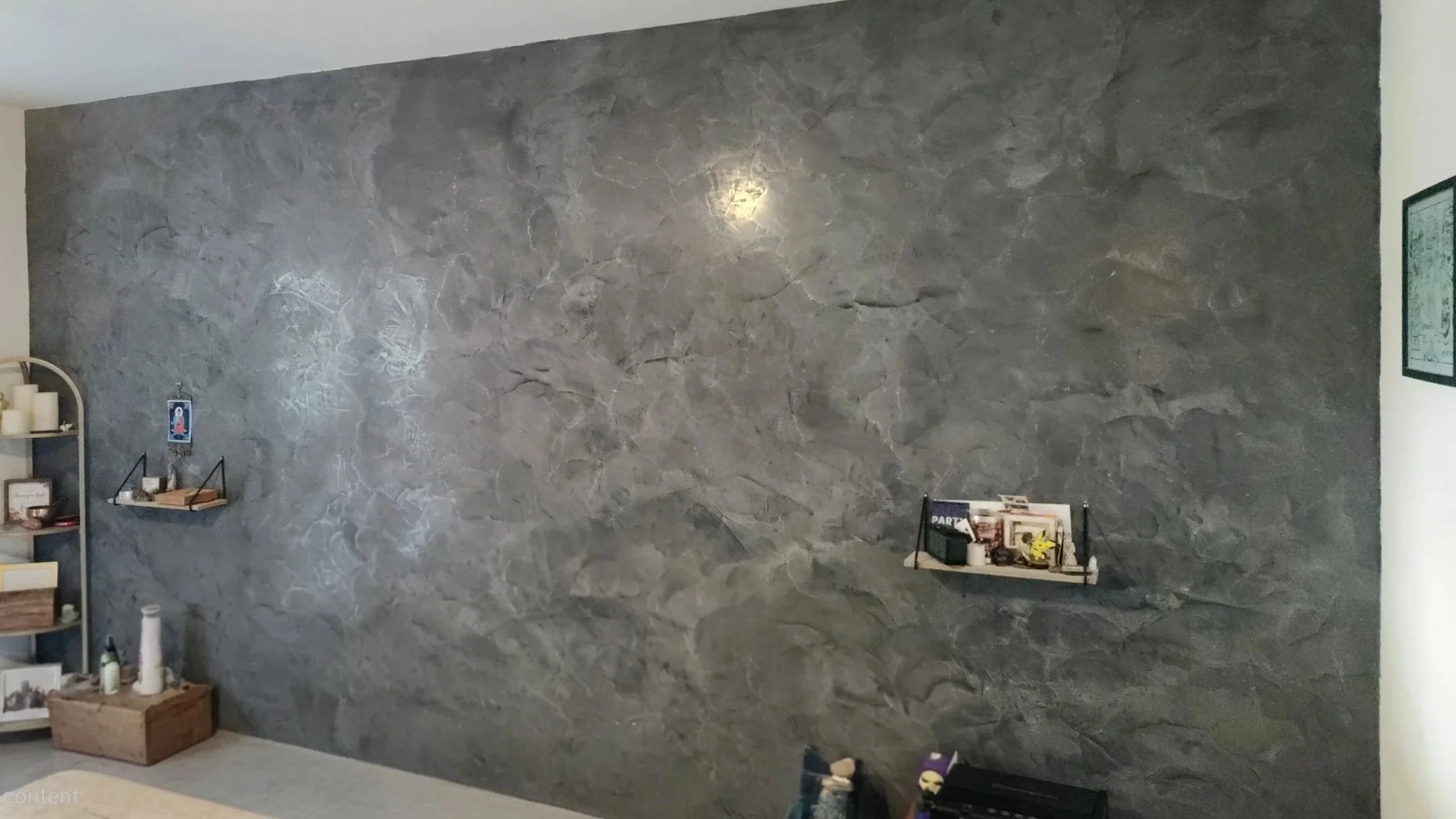 Interior view of a room featuring a large textured gray stone wall with a wall-mounted shelf holding books and decor. To the left, a corner shelf displays candles, framed photos, and decorative items. The floor is carpeted, and part of a bed or furniture is visible at the bottom of the image.