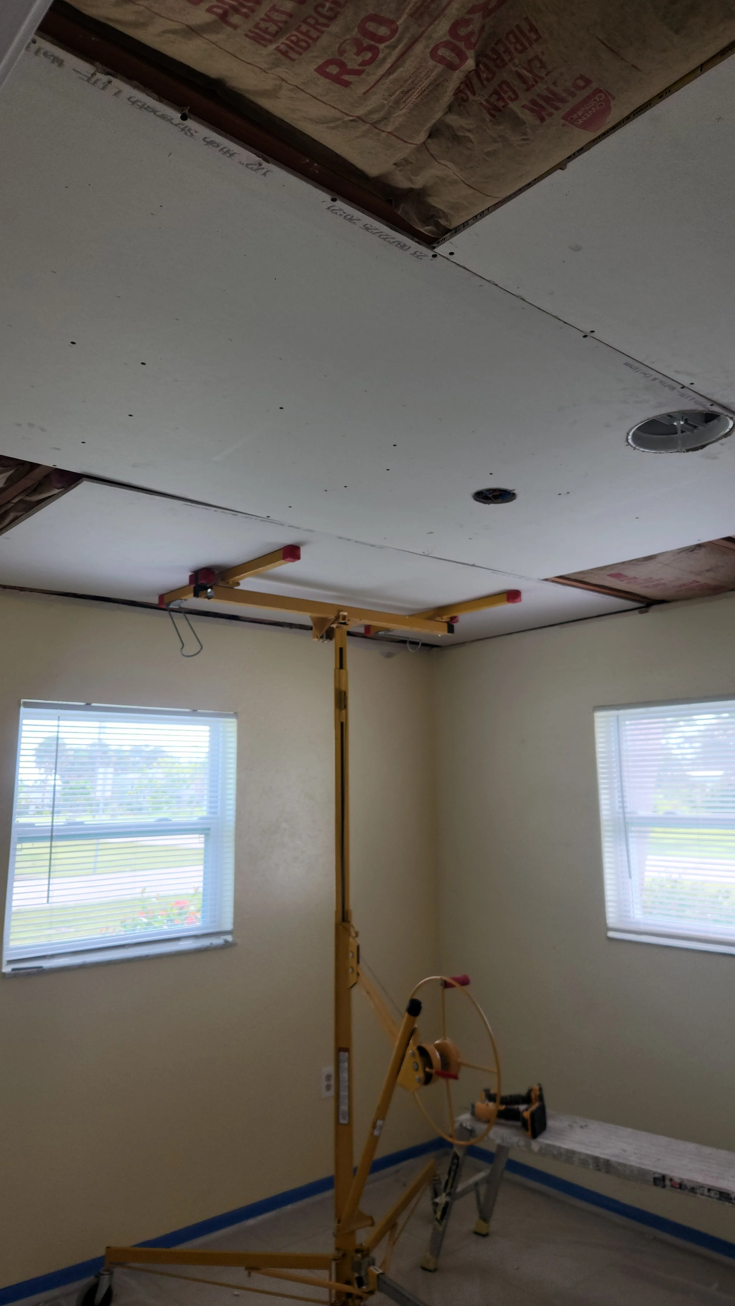Room under renovation with a lifted ceiling drywall, a lift, and two windows with blinds.