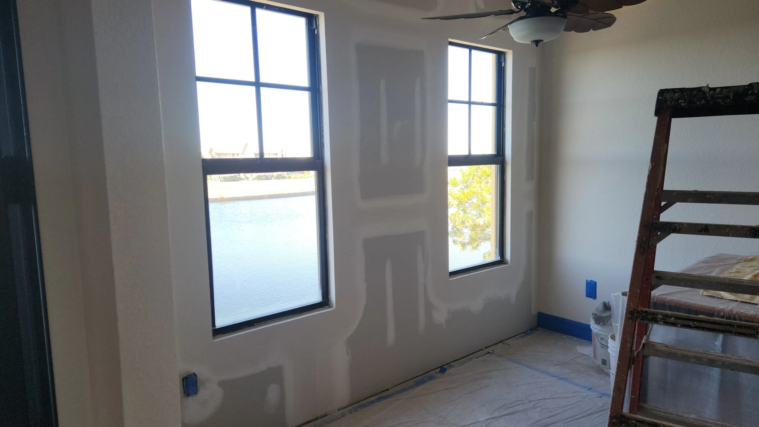 Room under renovation with two large windows overlooking water, unfinished drywall with visible joints, a ladder, and construction supplies.