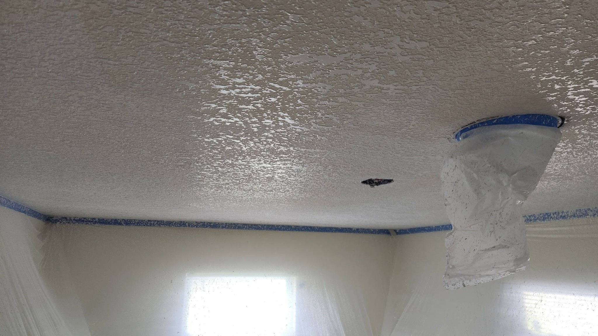 Ceiling being painted with white textured paint, painter tape around edges, and a plastic cover over a light fixture.