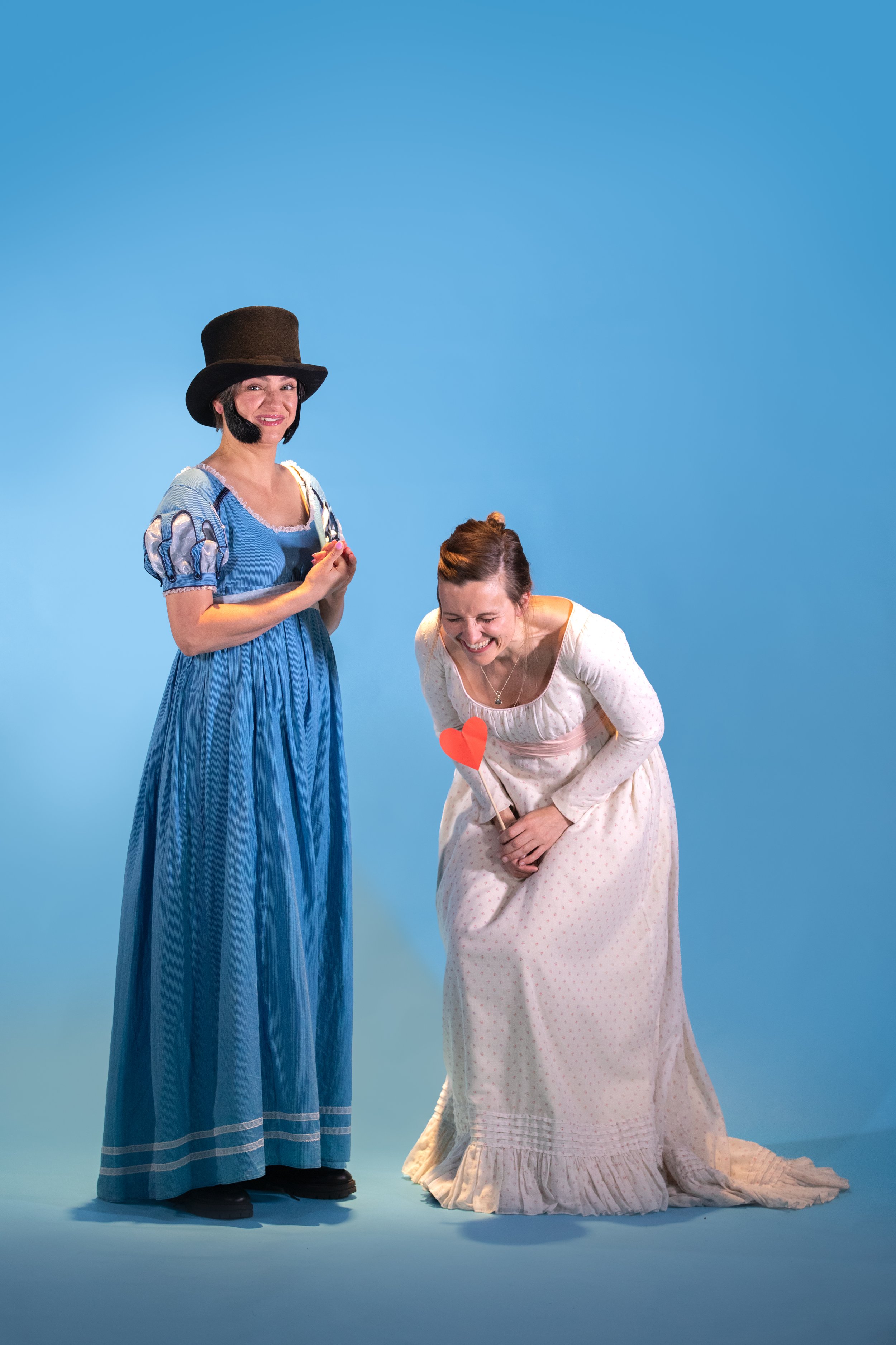 Two women in Regency dresses. One wearing a top hat with sideburns, one doubled over in laughter.