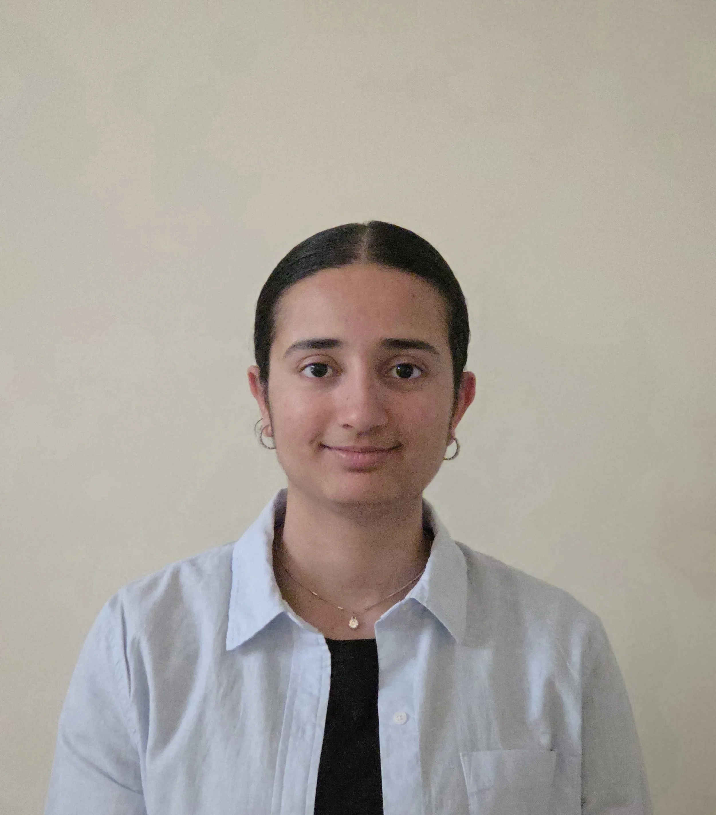 A woman with dark hair tied back, wearing a white shirt, black top, and gold hoop earrings, standing against a plain beige wall.