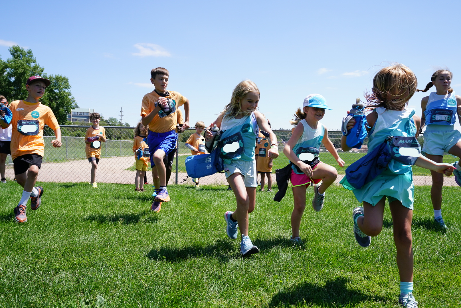 Children participating in a running event outdoors on a sunny day, running on a grassy field near a chain-link fence with a soccer field in the background.