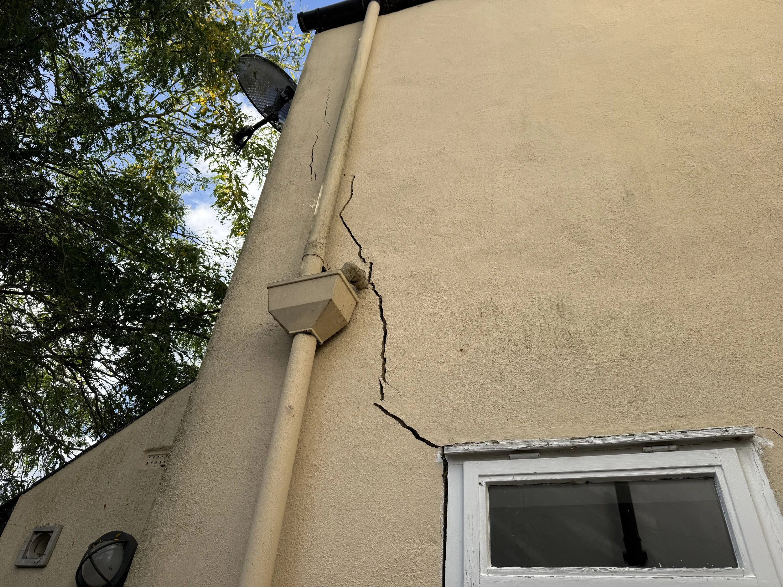 The image shows the corner of a beige house exterior with a vertical crack in the wall. There is an outdoor electrical box and a white window frame at the bottom right. A round outdoor light fixture is mounted near the electrical box. A rain gutter pipe runs vertically along the wall. In the background, there are green trees and a partly cloudy sky.