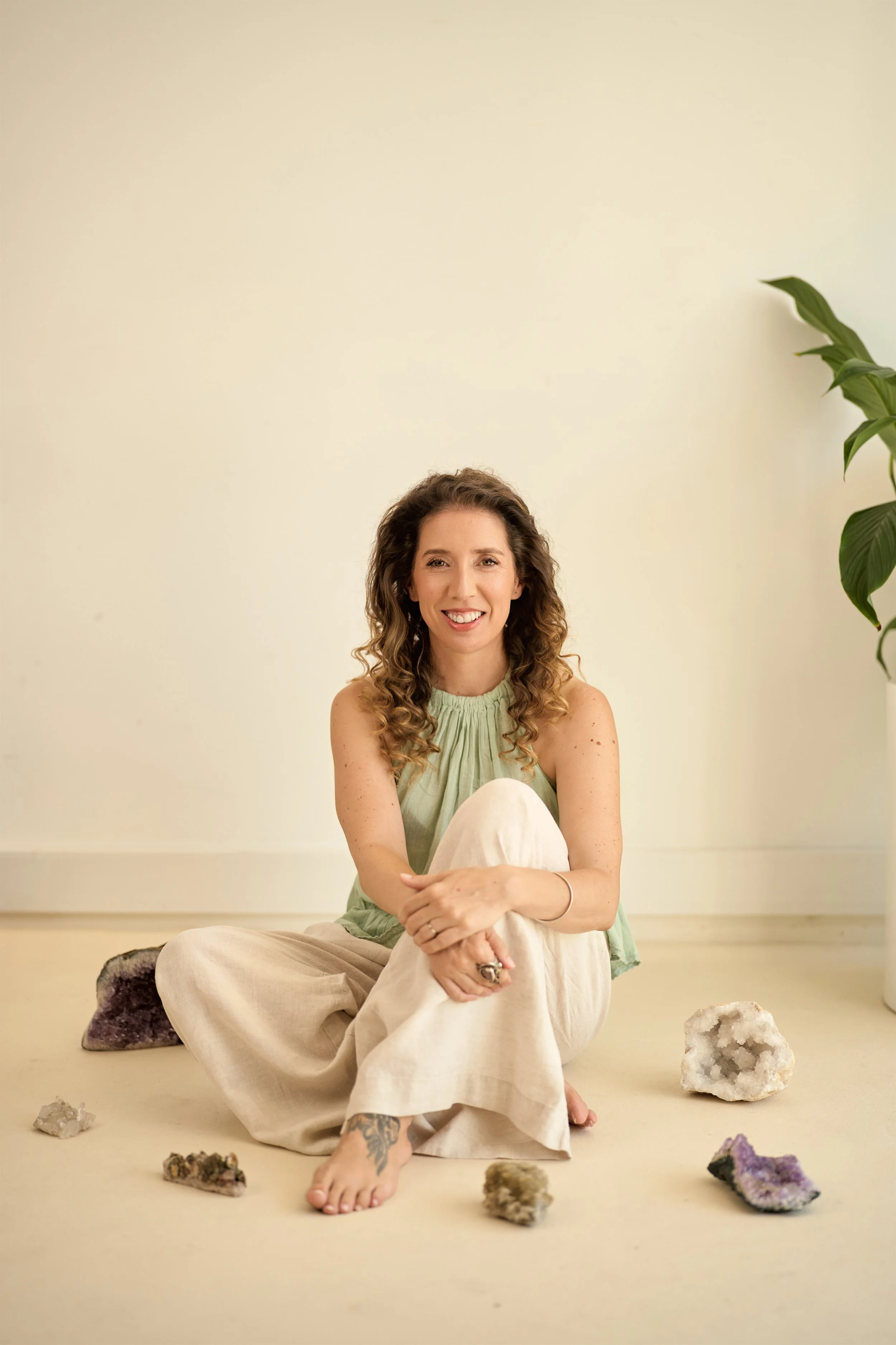 Mulher sentada no chão, sorrindo, rodeada por cristais e pedras, com uma planta ao lado, em um ambiente de fundo branco.