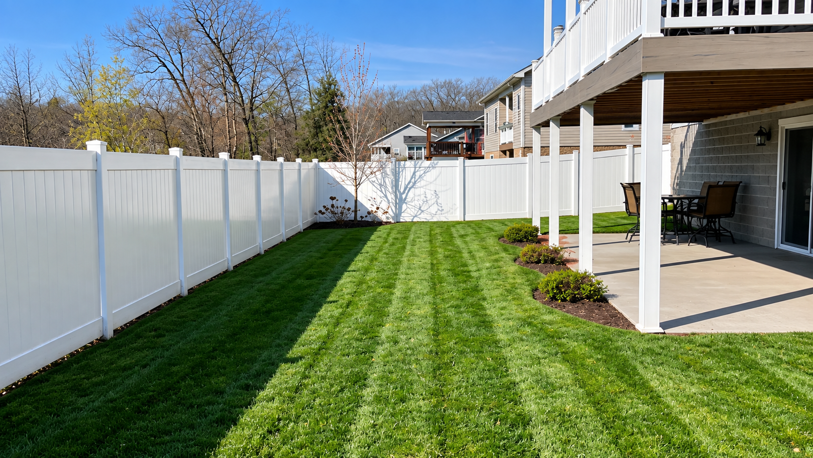 Clean backyard with green grass, white vinyl fence, small trees, and a covered patio with outdoor furniture.