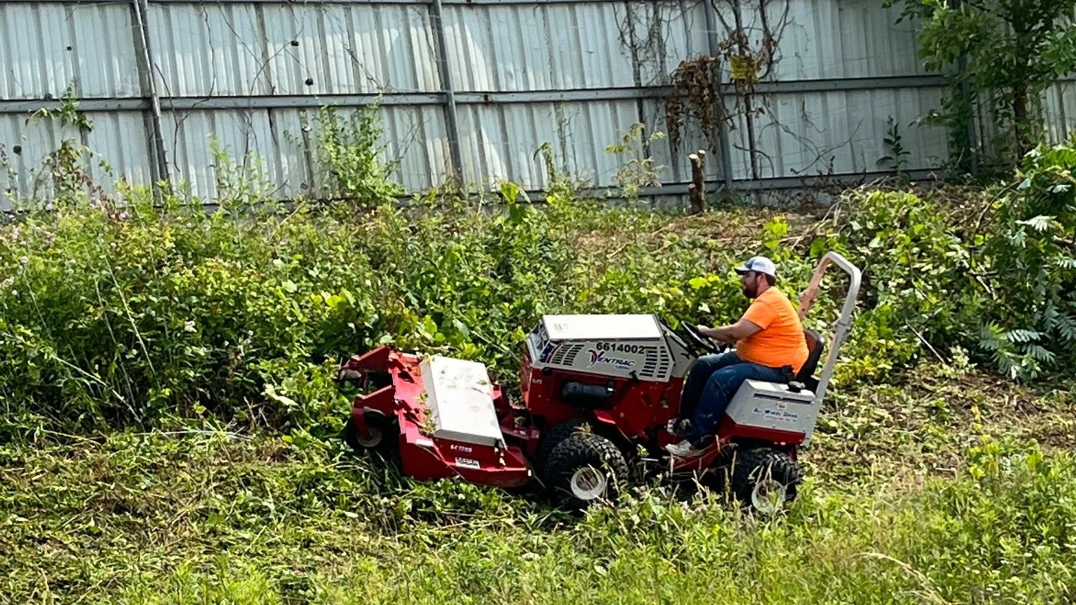 A man in an orange shirt and white cap operates a red and white riding mower in a grassy, overgrown backyard with a metal fence in the background.