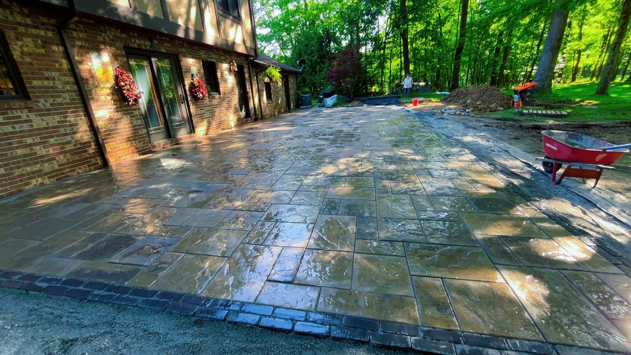 A newly paved outdoor patio with stone tiles in front of a brick house with decorated windows. Construction tools and dirt piles indicate ongoing work, and a person is seen in the background.