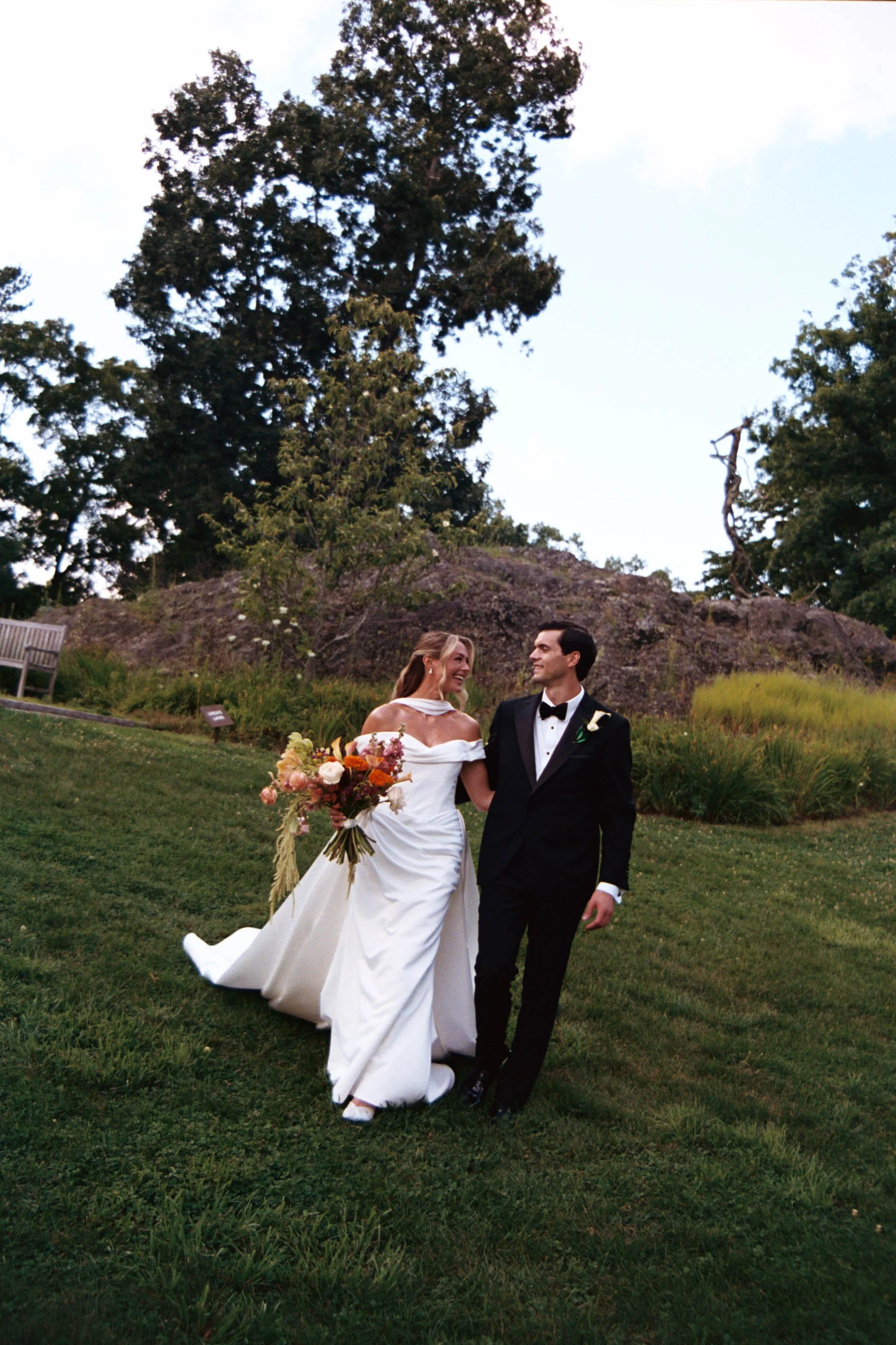 A bride and groom walking outside on a grassy lawn during their wedding, surrounded by trees and rocks, with the bride holding a bouquet of flowers. Boston wedding venue with chic film photos