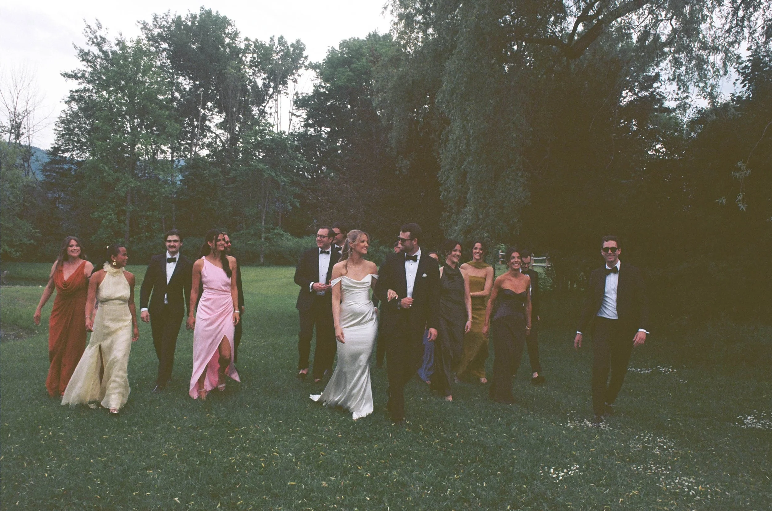 Wedding party in formal black tie attire walking together outdoors on a grassy area with trees in the background,  at a wedding in Manchester Vermont. Wedding bridal party on 35 mm film.