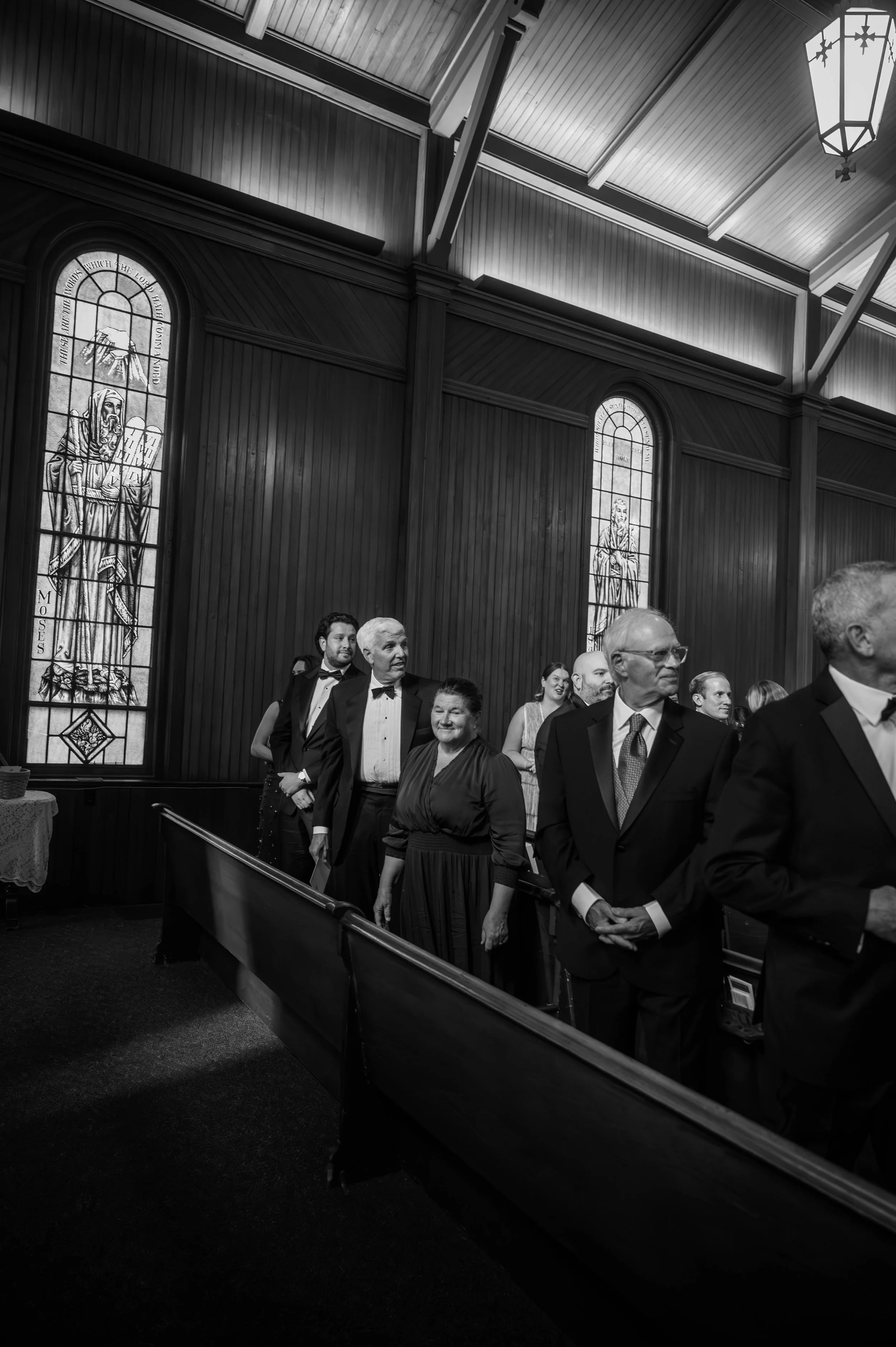 People dressed in formal attire standing in line inside a church wedding, with stained glass windows and wooden paneling in the background in Manchester Vermont.