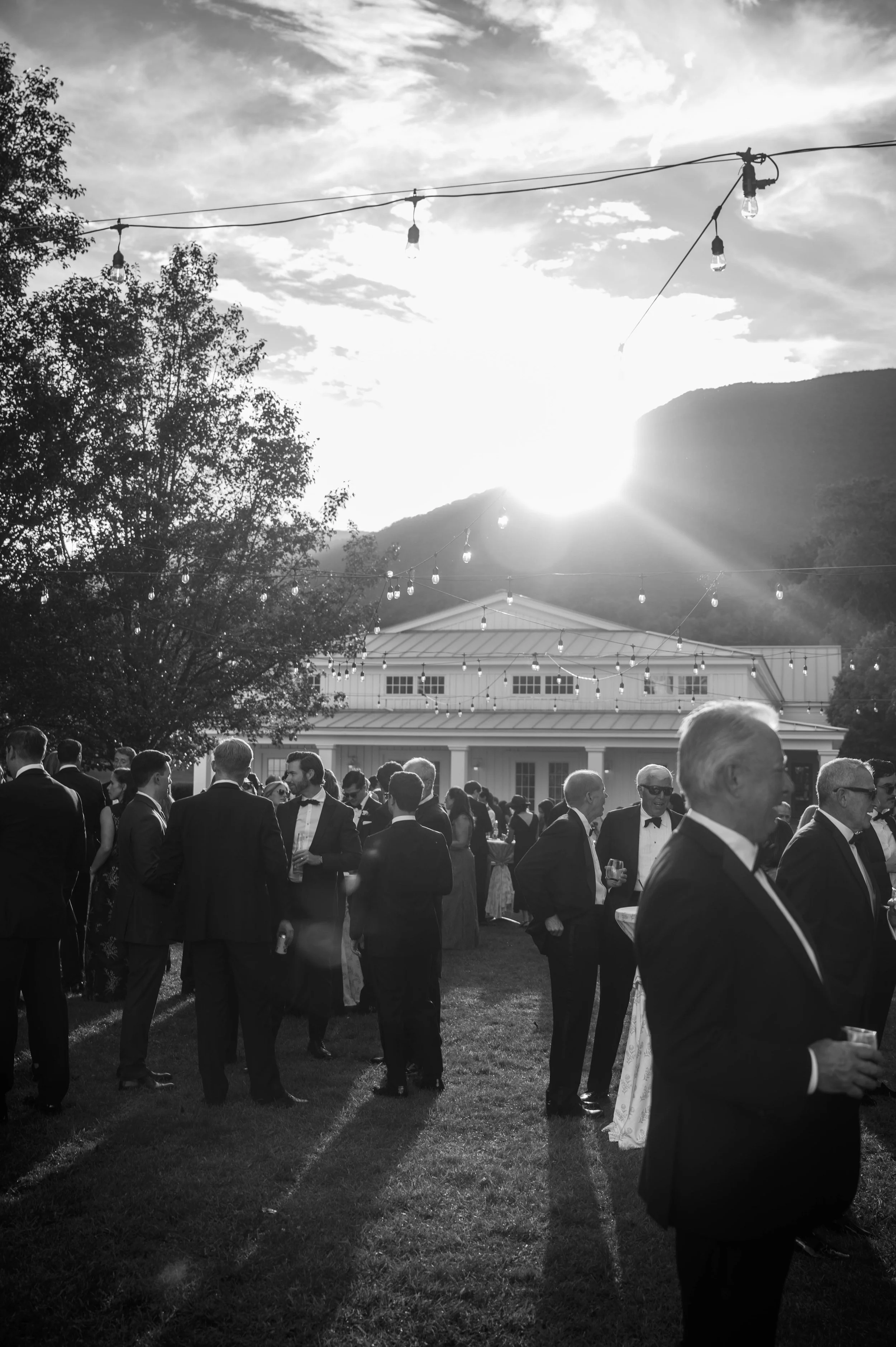 Black and white photo of a group of people in formal attire at an outdoor wedding during sunset, with string lights overhead and a building in the background at the equinox inn and resort.