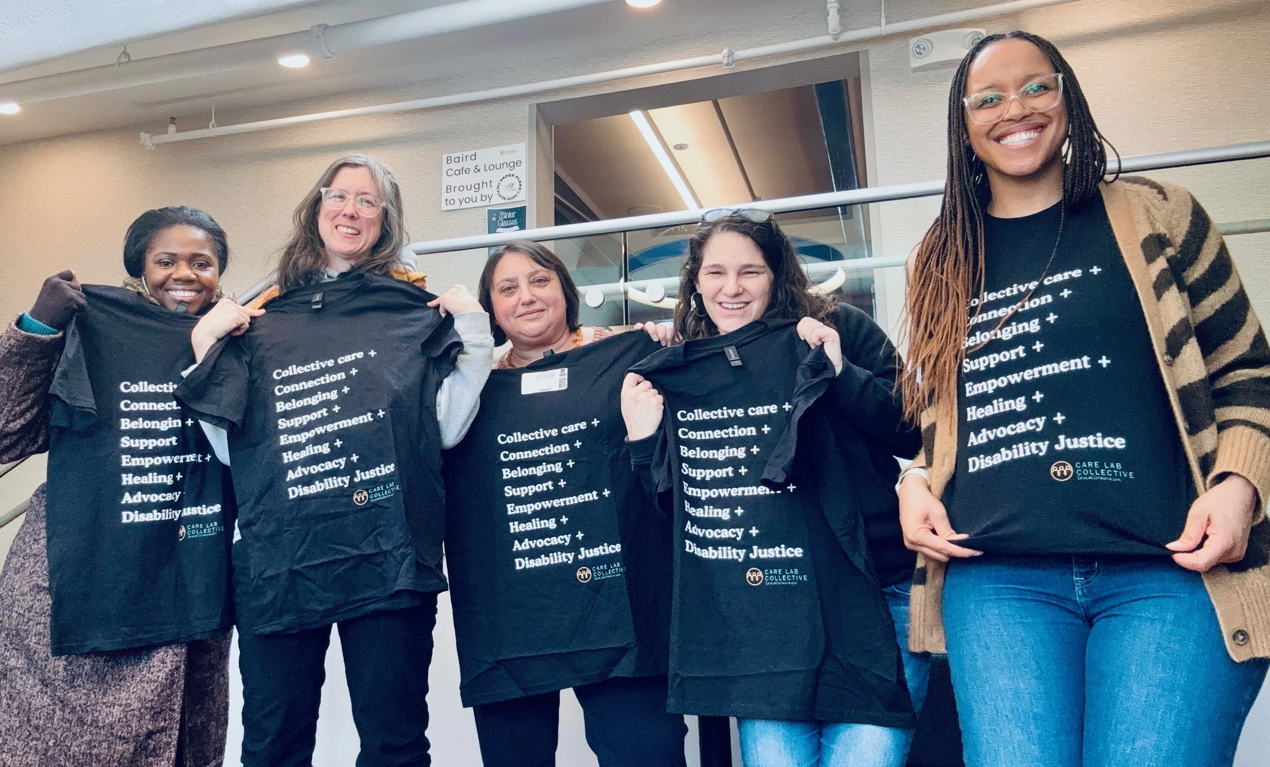 Five women smiling and holding black T-shirts with white text. The T-shirts have messages about collective care, connection, belonging, support, empowerment, healing, advocacy, and disability justice. They are indoors, standing in a line, with a sign and reflective glass window in the background.