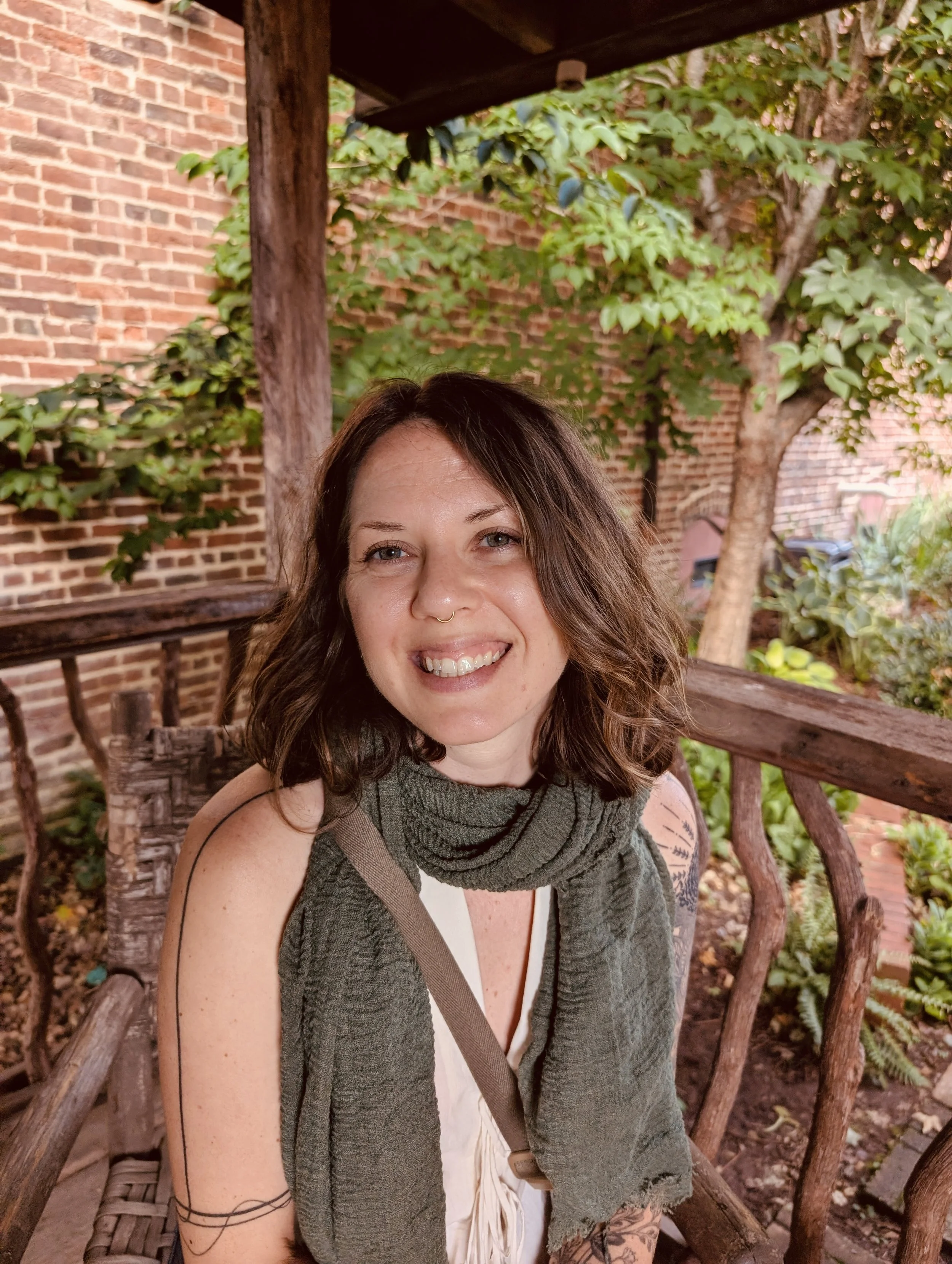 A woman with brown curly hair smiling, wearing a white top, a gray sleeveless sweater, and tattoos on her arm, sitting outdoors on a wooden bench with green plants, trees, and a brick building in the background.
