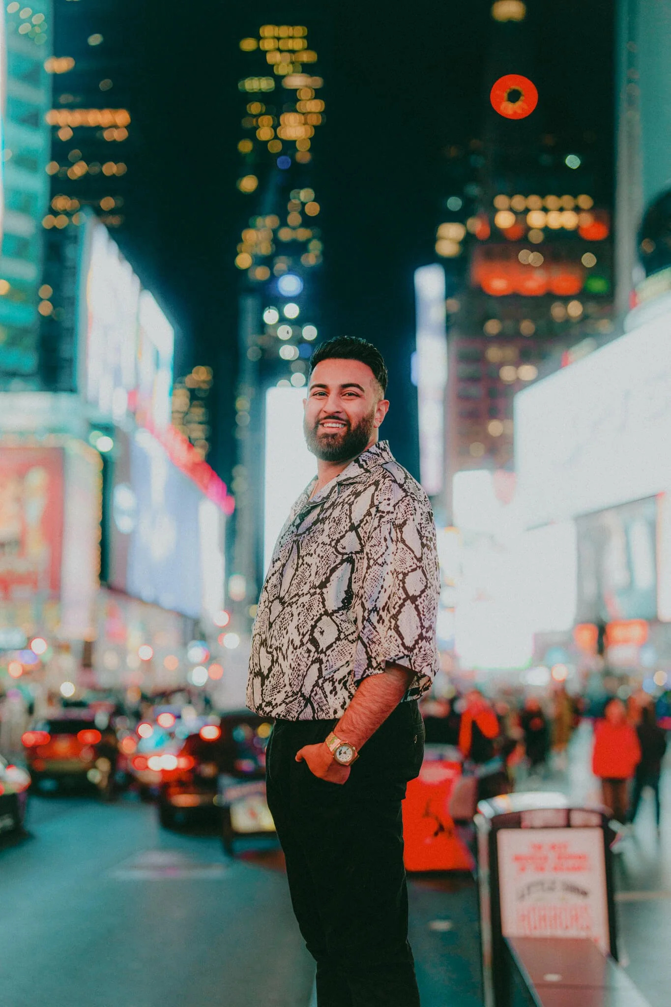 A smiling man with dark hair and a beard stands in Times Square at night, wearing a snake print shirt and black pants, with bright city lights and billboards in the background.