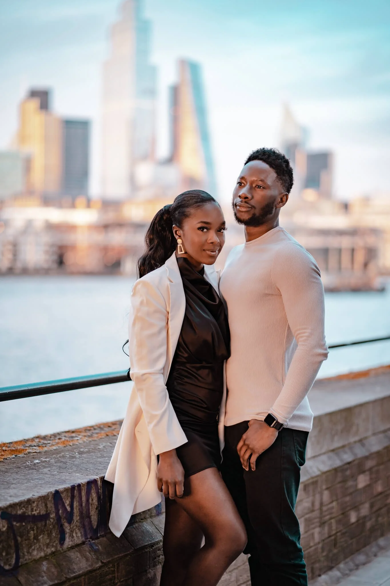 A fashionable Black man and woman stand close together by a waterfront with a city skyline in the background during daytime. The woman wears a black dress and white blazer, and the man wears a white long-sleeve shirt and black pants.