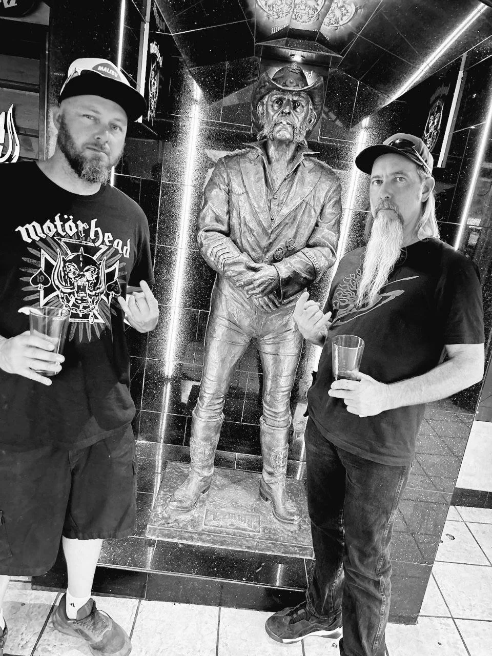 Two men with beards and caps standing on either side of a cowboy statue inside a building with dark tiled walls, holding drinks