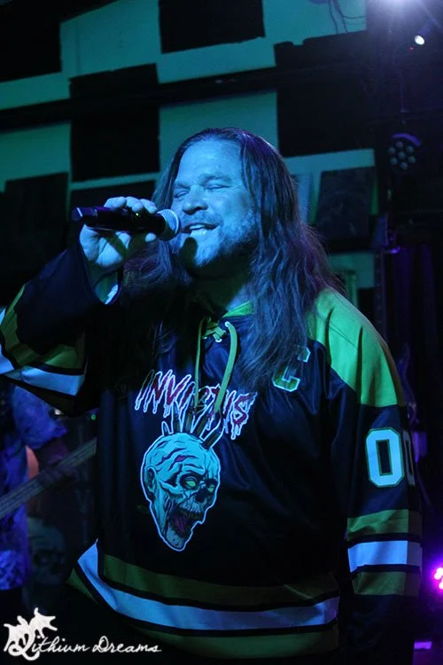 A man with long hair singing into a microphone, wearing a dark hockey-style jersey with green, yellow, and black colors and a graphic of a zombie's head on the front, in a dimly lit venue.