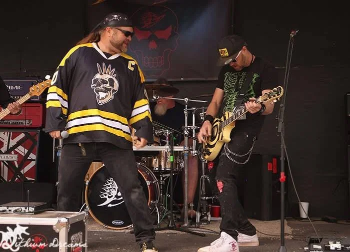 Two musicians performing on stage, one with a black hockey jersey with a skull logo, the other with black clothing and a cap, both playing guitars, with a drum set behind.