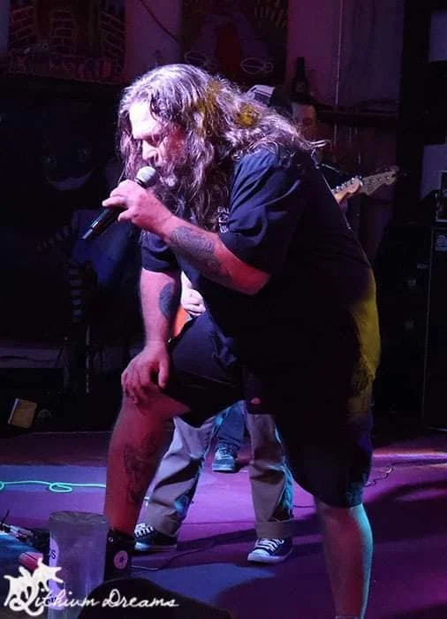 A man with long hair and tattoos singing into a microphone on stage, with one foot on a monitor speaker, in a dimly lit venue.