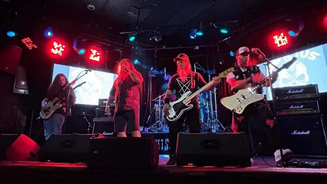 A four-member band performing on stage with instruments, including guitars and drums, inside a club with a red and black color scheme and stage lighting.
