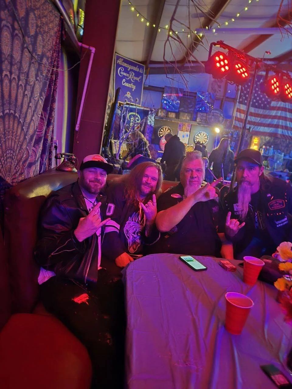 Four men sitting at a table in a lively bar or club, making rock and roll hand gestures. The setting is decorated with string lights, American flag, and darts in the background.