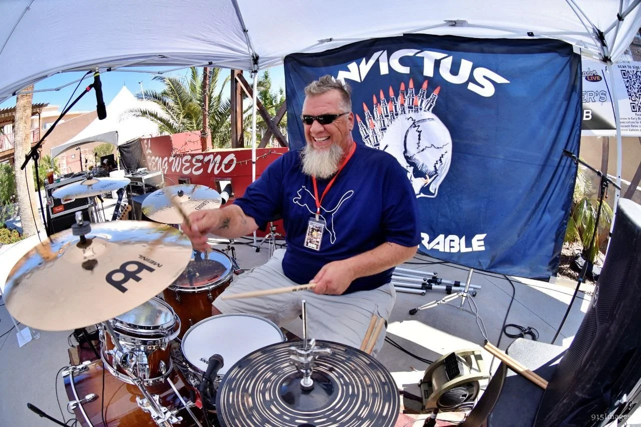 A man with sunglasses and a beard playing the drums outdoors under a white canopy, smiling. There are palm trees and buildings in the background. He is wearing a blue shirt with a Puma logo and a lanyard. Behind him, a large flag with the words 'Invi