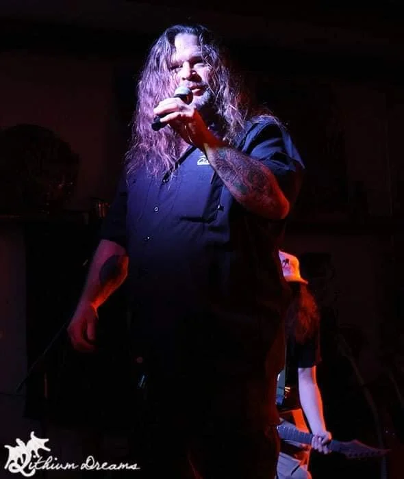 A male singer with long, wavy hair, wearing a dark shirt, performing with a microphone on stage in a dimly lit venue.