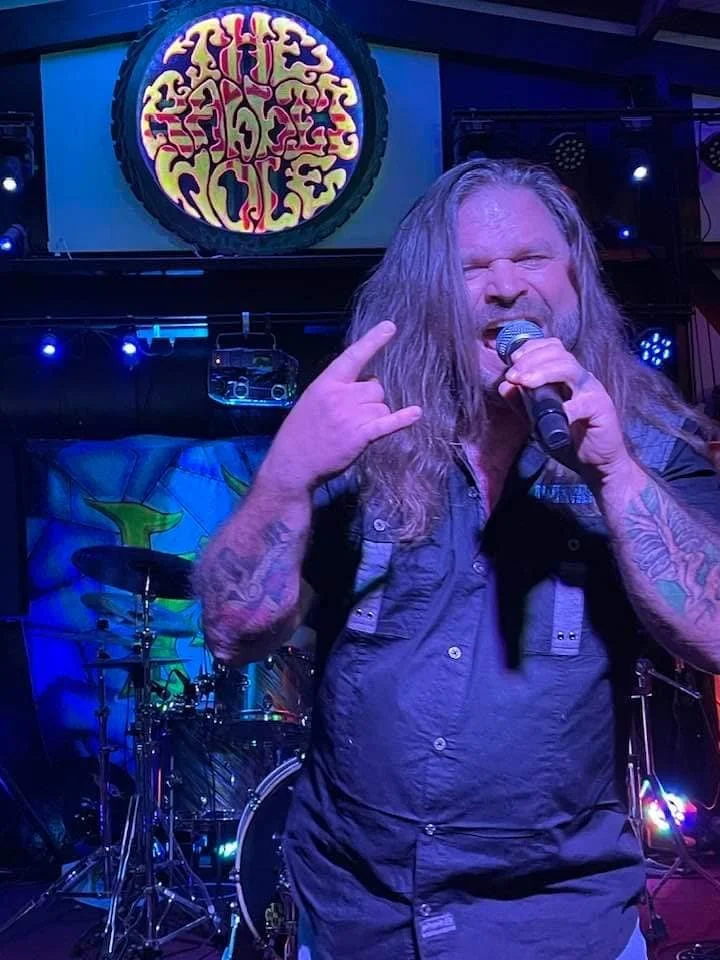 A long-haired man with tattoos on his arms singing into a microphone on stage at a rock concert, making a hand gesture with his left hand. There are drums and colorful background artwork behind him.