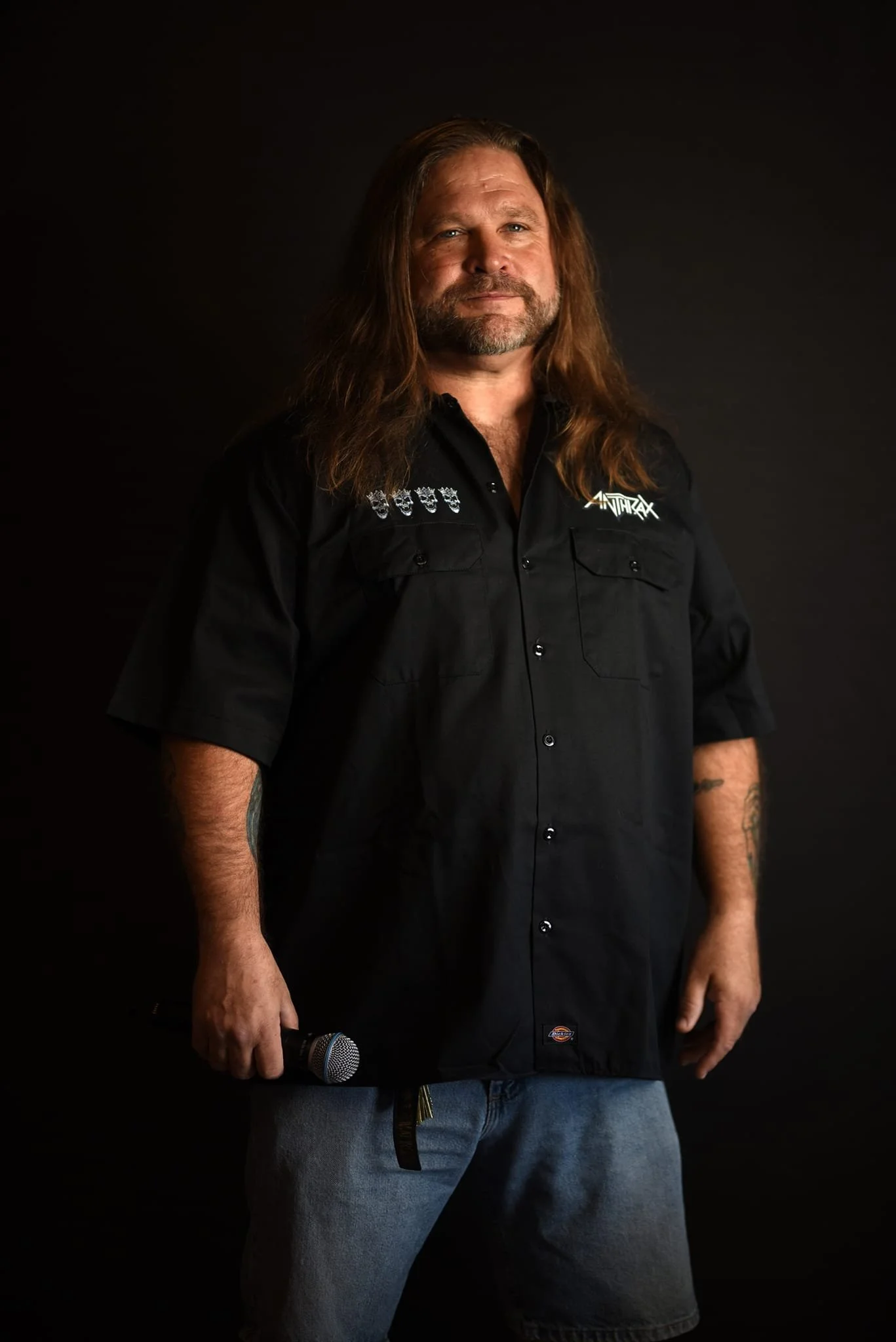 A man with long hair and a beard holding a microphone, wearing a black shirt with patches, standing against a dark background.