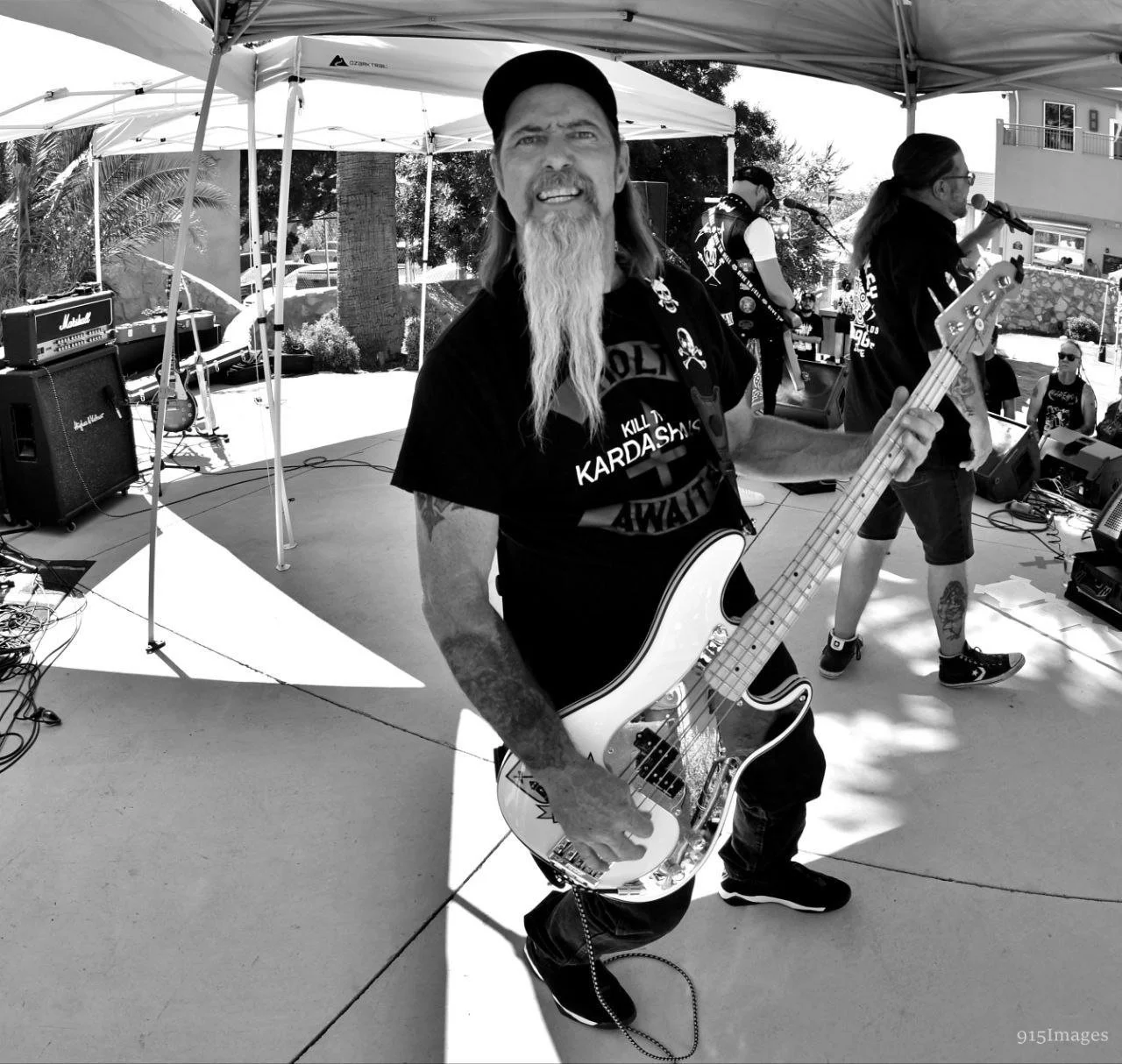 A man with a long beard and a backwards cap playing bass guitar on an outdoor stage with a band.