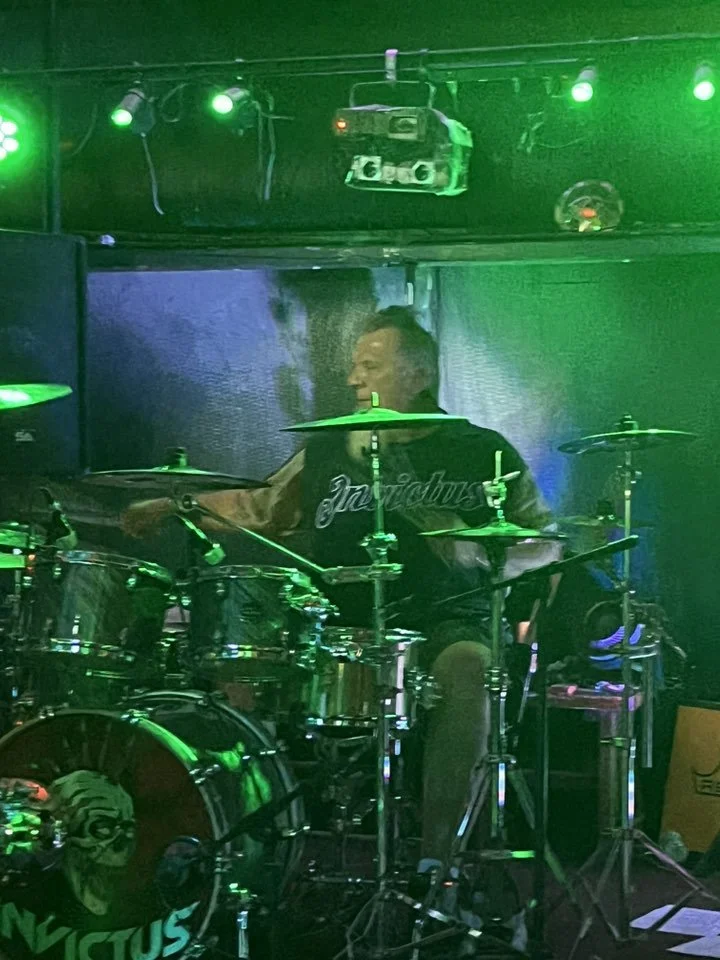 A person playing a drum set on a stage illuminated by green lighting, with stage equipment and a backdrop visible.