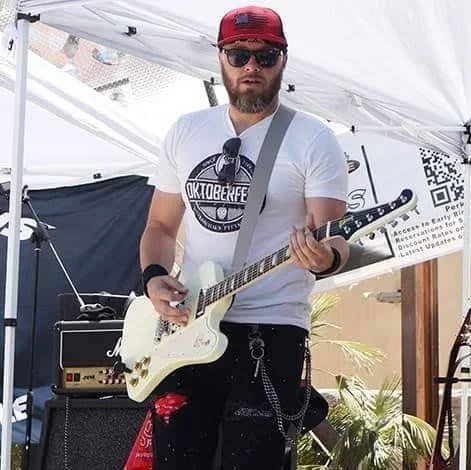 Man with beard wearing red cap, sunglasses, and white T-shirt playing electric guitar at outdoor event