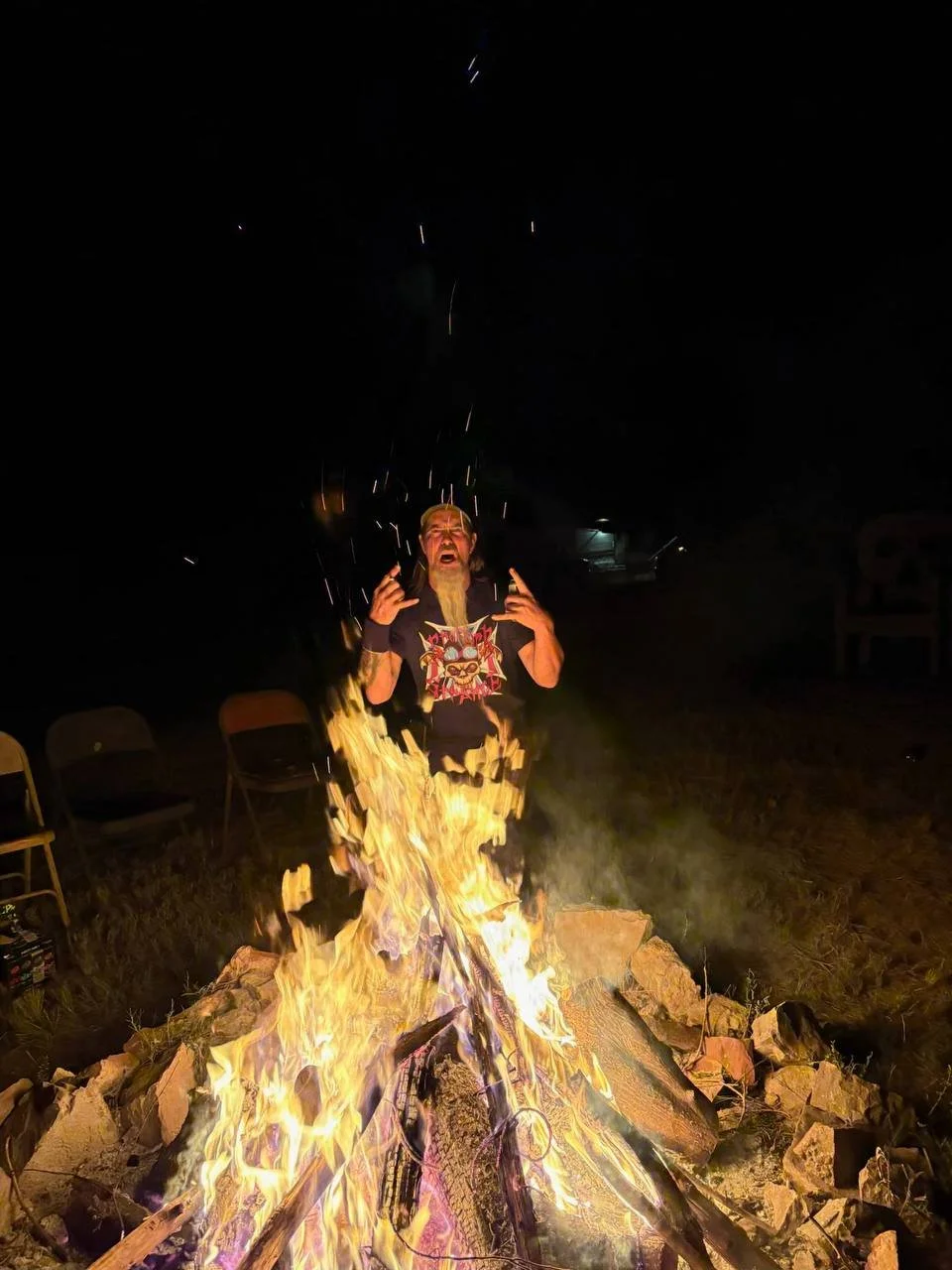 A man with a beard and glasses standing near a large bonfire at night, with chairs in the background.