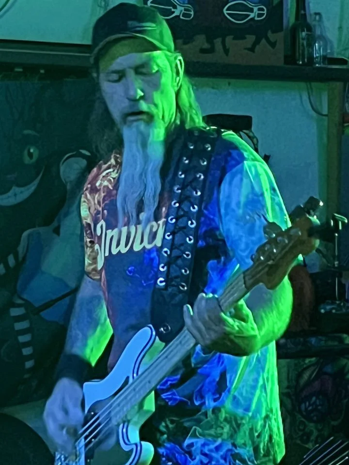 Musician with long hair, glasses, and a beard playing a guitar in a dimly lit room with blue and green lighting.