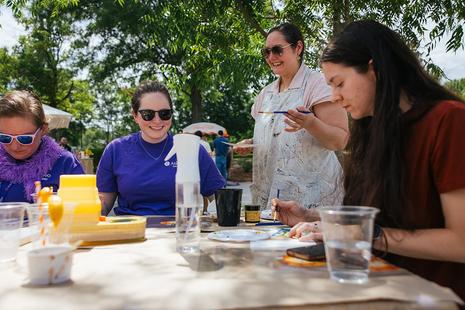 Four women painting with Stacia Patin from Stacia's Studio, engaging in a watercolor painting art activity with paints and brushes, surrounded by trees on a sunny day.
