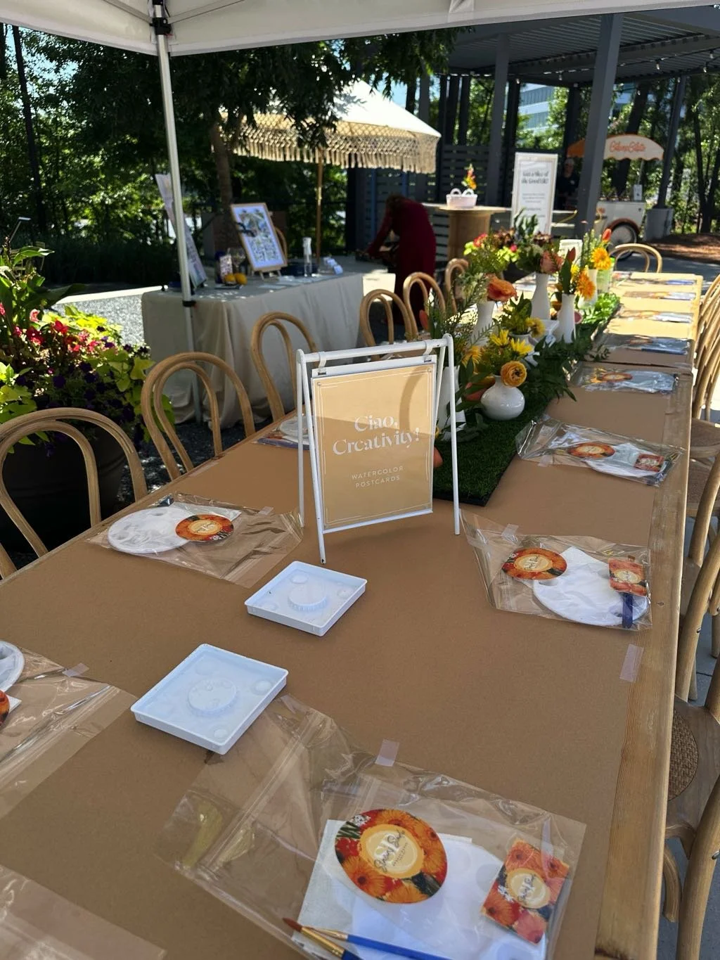 A long outdoor table decorated with a floral centerpiece and place settings with paper napkins and water color postcards for a creative event or gathering. There is a sign that reads "Ciao, Creativity! Watercolor Postcards".