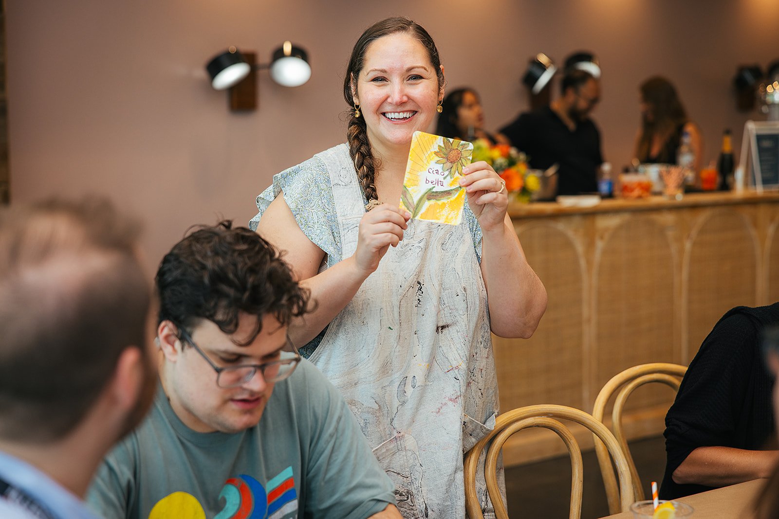 Stacia Patin, artist holding a watercolor painting of a sunflower and the words 'Ciao bella' at Stacia's Studio watercolor event.