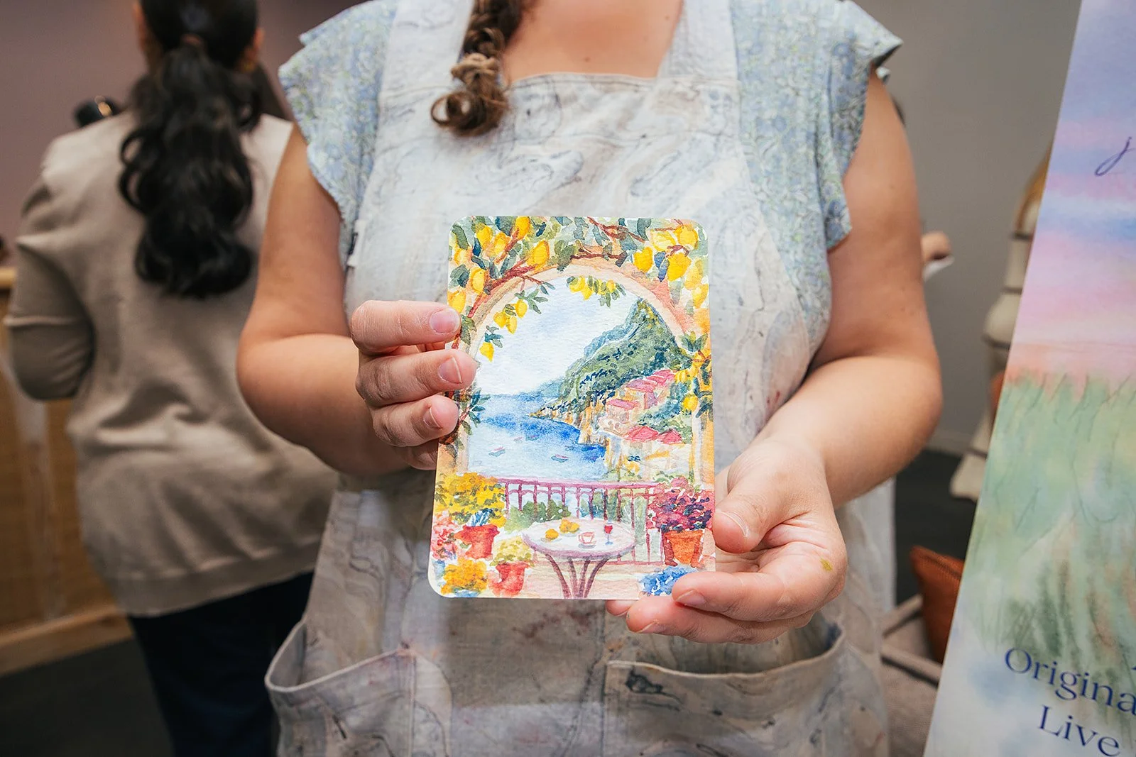 Person holding a colorful painting of a seaside landscape with a bridge, trees, and a terrace with a table and chairs.