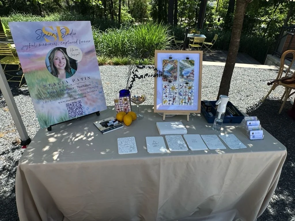 Outdoor booth with a sign for Stacia's Studio, featuring a photo of a woman and contact info, set up with watercolor paintings, lemons, a water spray, and various design materials under trees on a gravel surface.