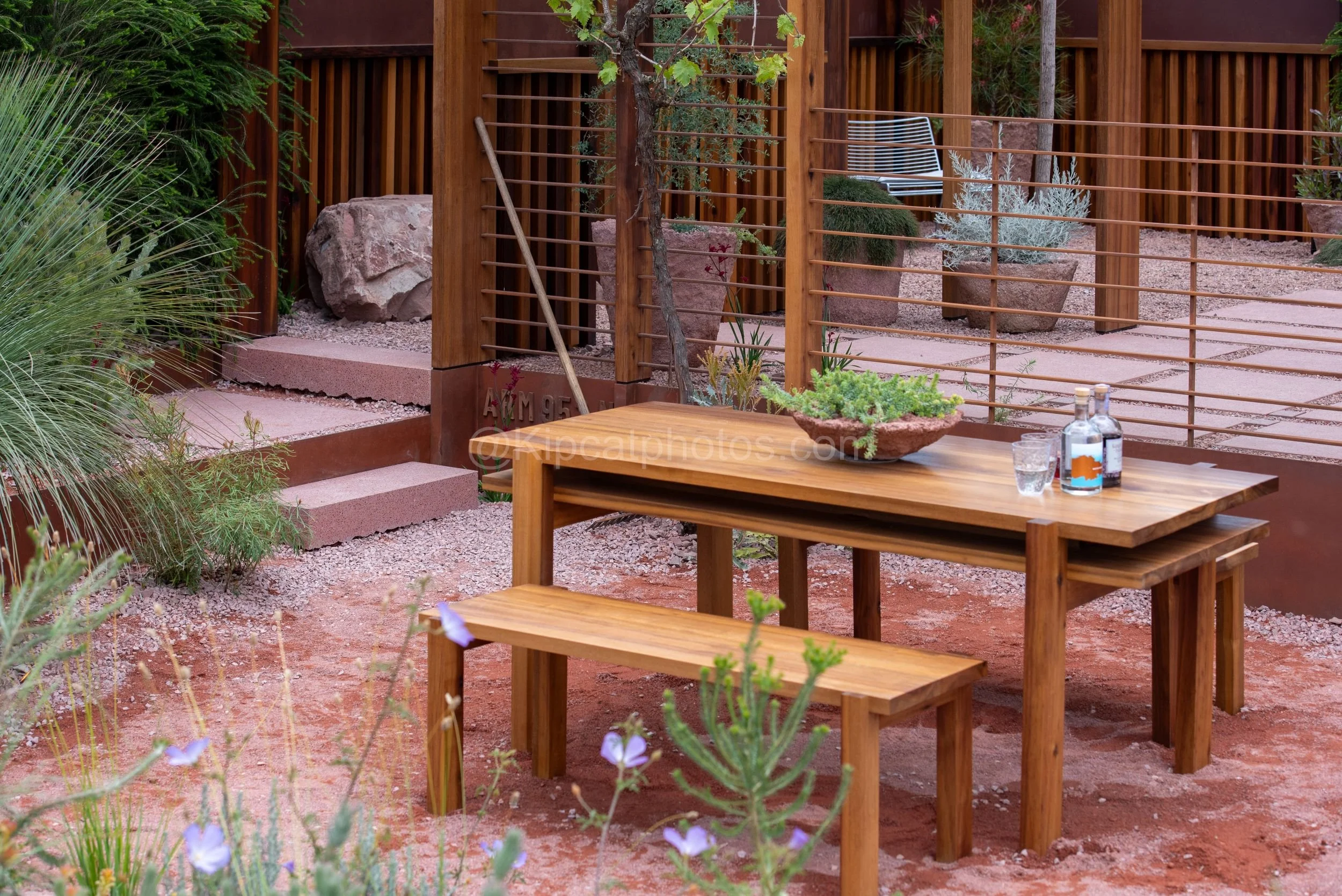 Timber dining table and benches in a red-earth Australian-style garden in the Max Parker-Smith garden at RHS Chelsea Flower Show 2026

A timber table and bench set stands on red desert-toned ground beneath a slatted timber pergola, with grass trees, 