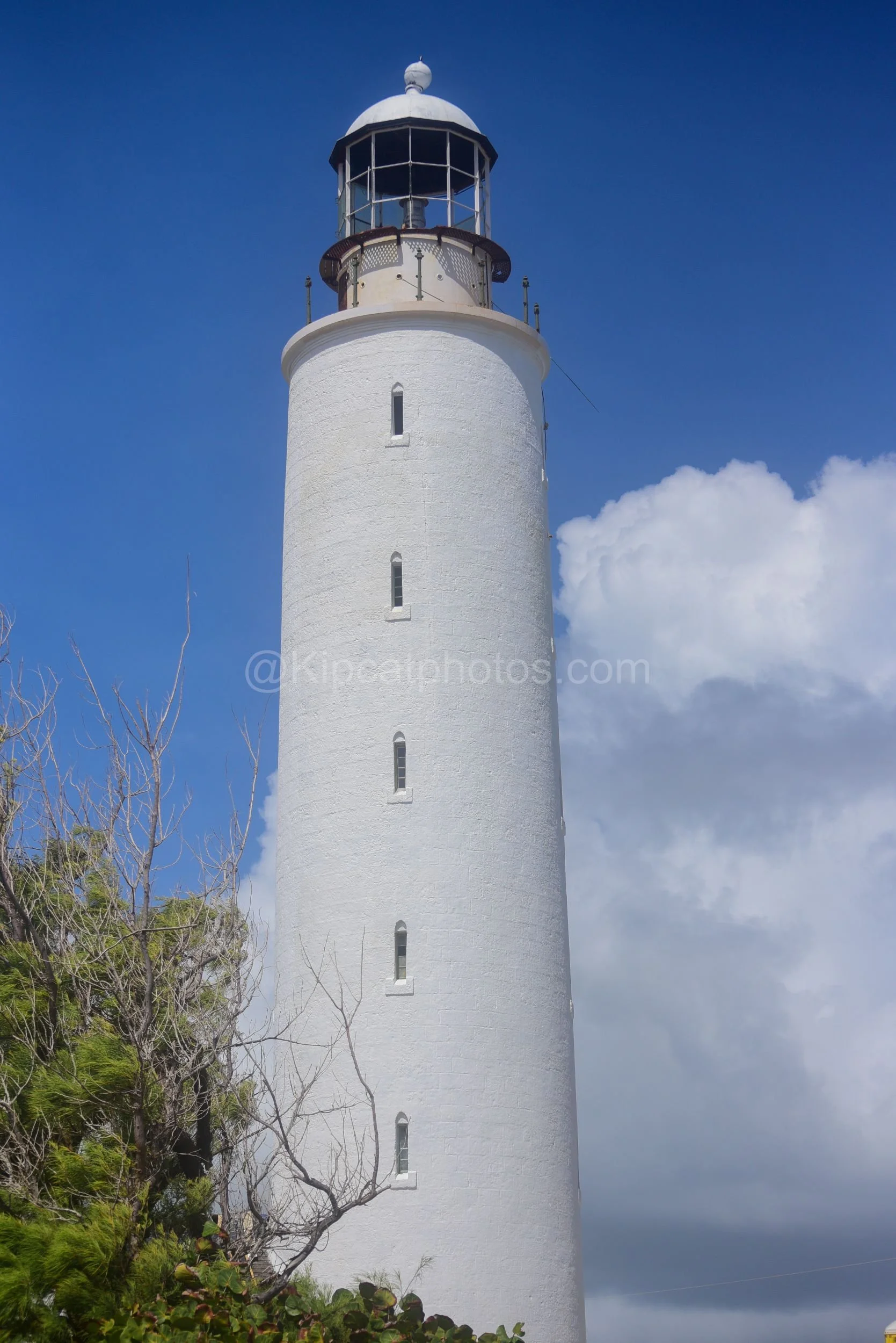 Ragged Point lighthouse tower in Barbados with its glazed lantern room and dome against a blue sky. Kipcatphotos.com

Bare branches frame Ragged Point lighthouse tower, its glazed lantern room and domed roof catching the sun against a backdrop of blu