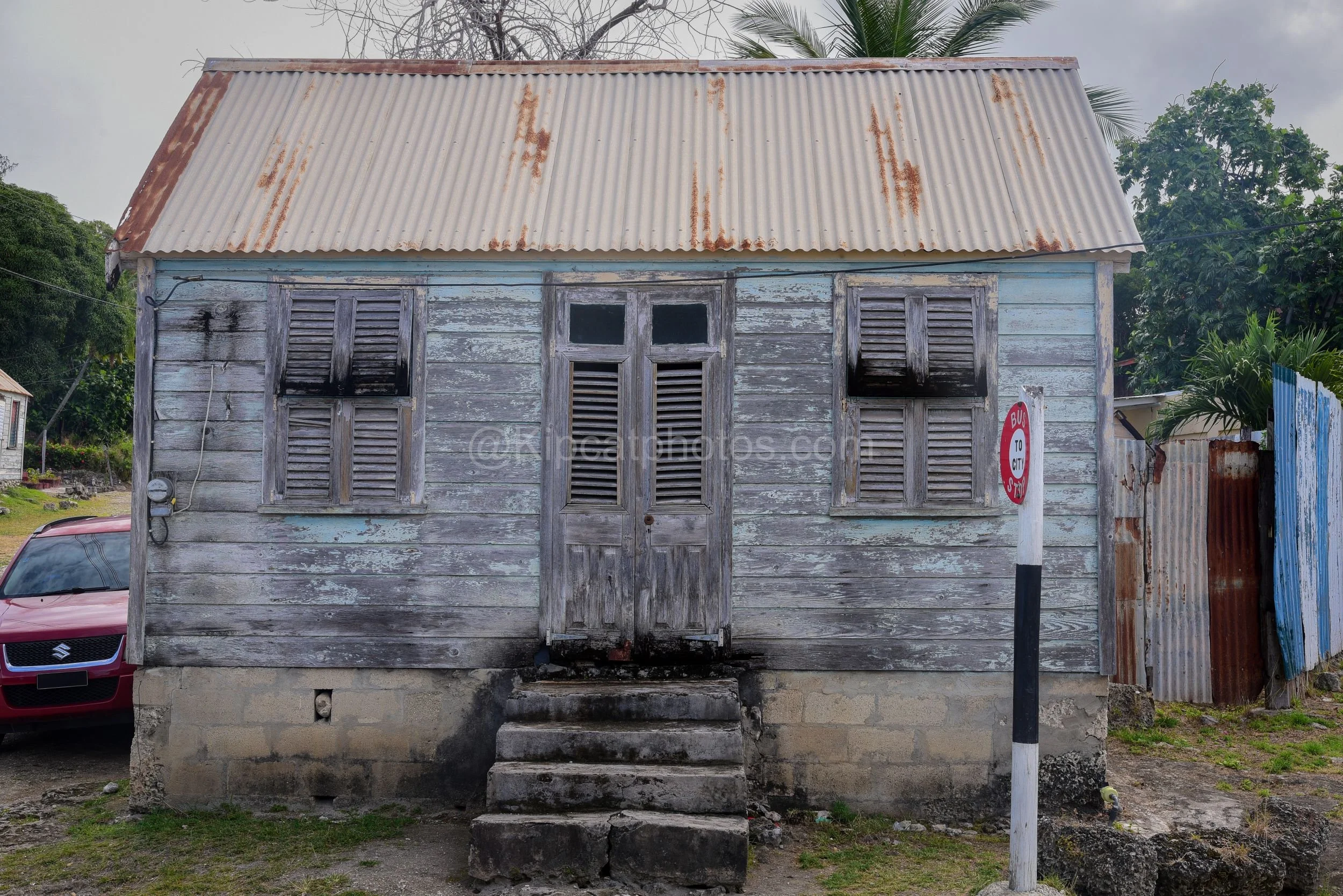A weathered blue-grey chattel house in Barbados beside a red car and a "Bus to City" stop sign. Kipcatphotos.com

This blue-grey chattel house has lost most of its paint to the sun, its shuttered windows and panelled door standing beside a rusted cor