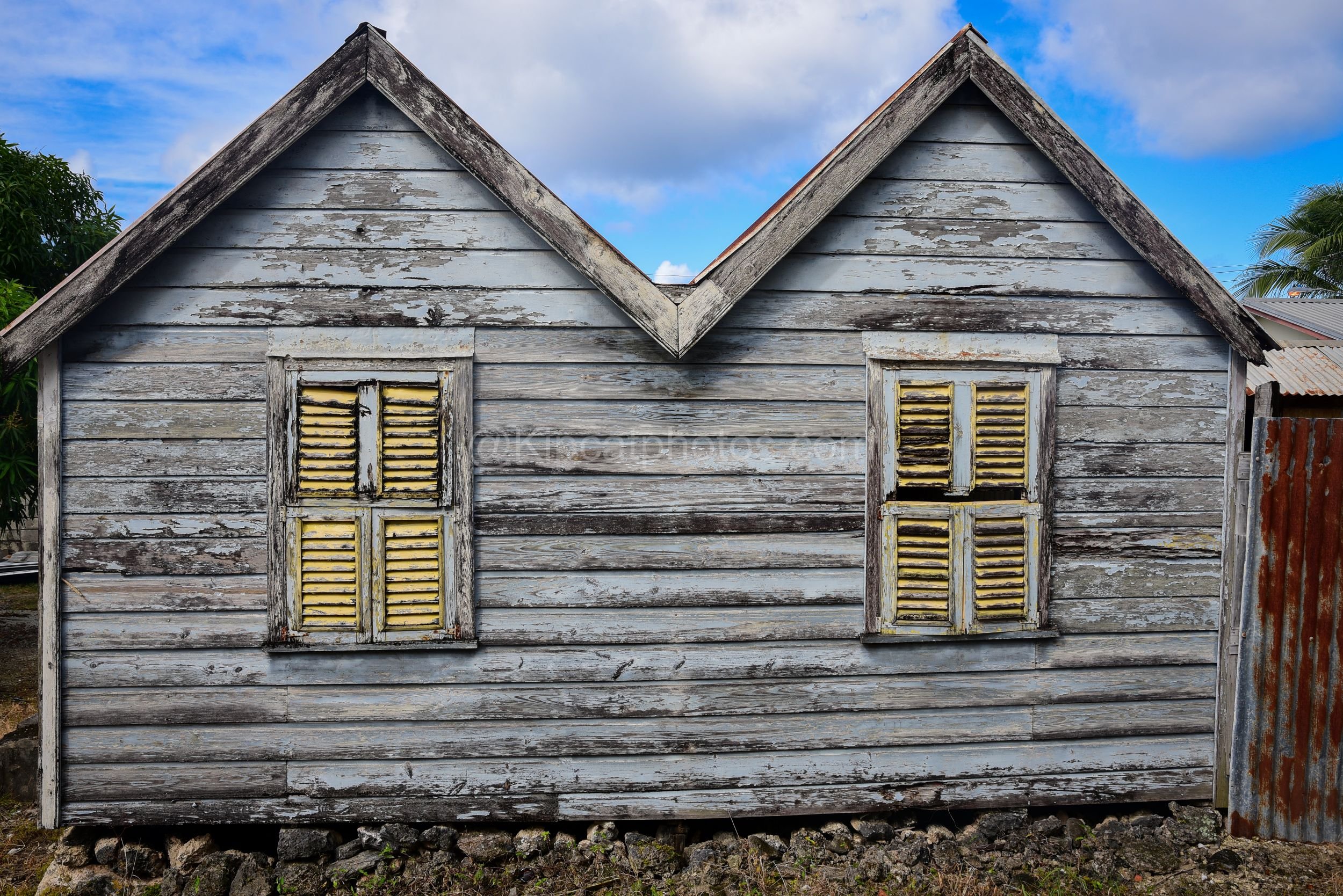 A weathered grey chattel house in Barbados with twin gables and yellow louvred window shutters. Kipcatphotos.com

Two steep gables meet at a central valley on this grey, sun-bleached chattel house, its yellow louvred shutters providing a splash of co